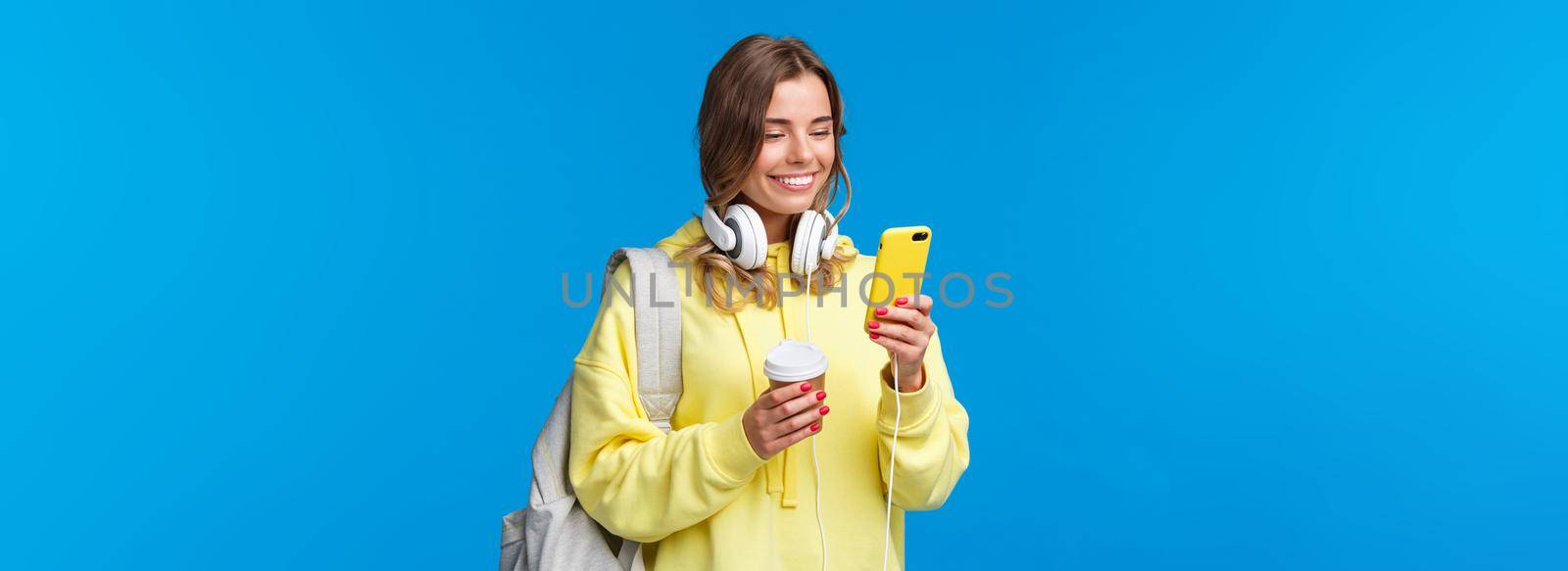 Girl using mobile phone to contact friend after college, texting as walking along street with backpack, take-away coffee cup and headphones, smiling joyfully smartphone display by Benzoix