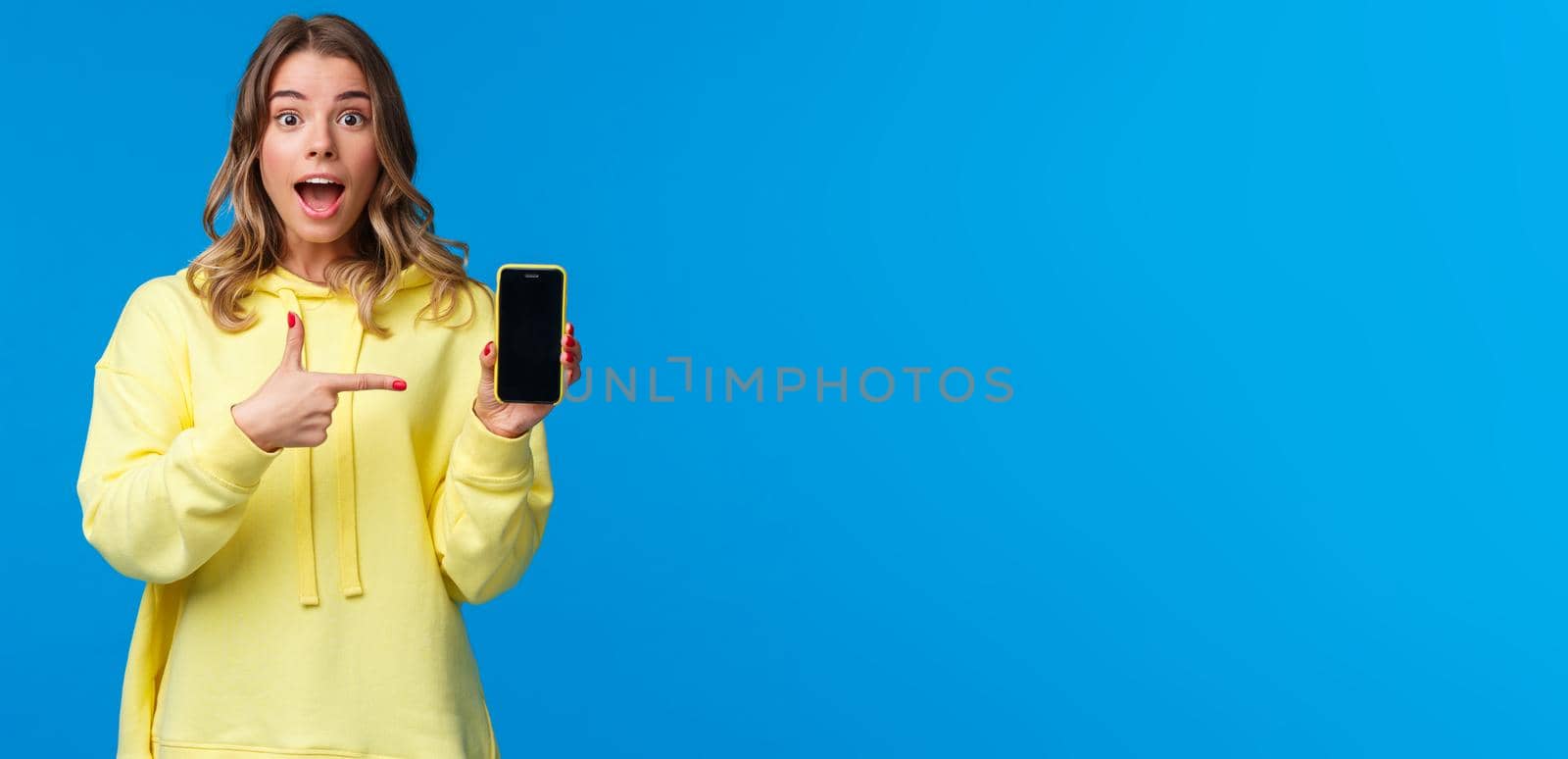 Impressed happy cheerful blond girl explain something as pointing mobile phone display, talking about new application or sharing her vacation photos, hold smartphone, look camera by Benzoix