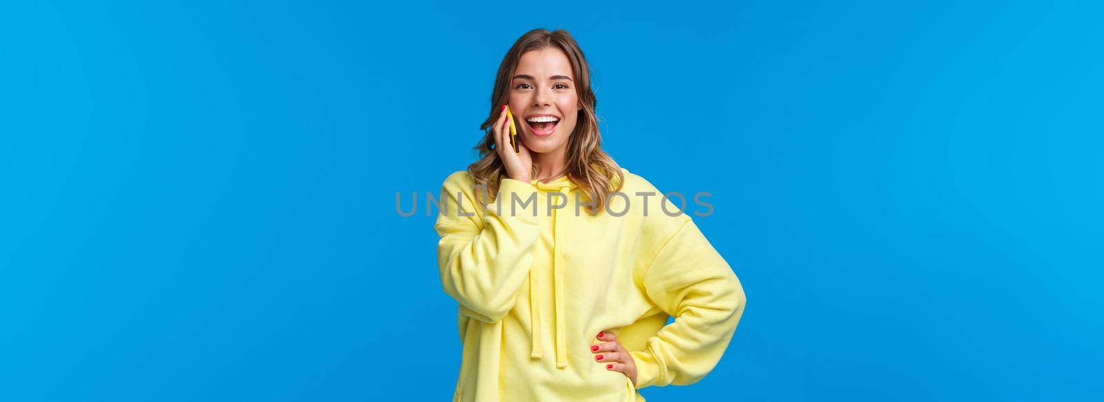Carefree good-looking european blond girl in yellow hoodie lively talking on mobile phone, smiling camera as booking table for friends in bar, order food delivery, blue background by Benzoix