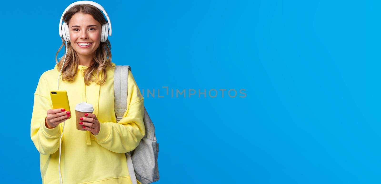 Education, lifestyle and teenagers concept. Cheerful young blond girl in headphones listening music and texting friends while waiting for them near cafe, drinking take-away coffee hold backpack.