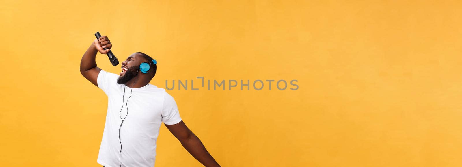 Portrait of cheerful positive chic. handsome african man holding microphone and having headphones on head listening music singing song enjoying weekend vacation isolated on yellow background.