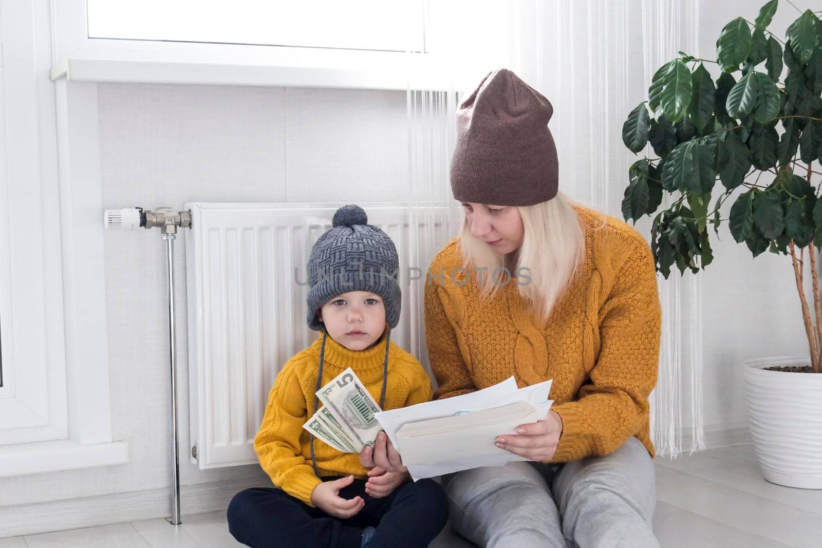 .A mother with a small child in a yellow sweater and hats is counting money and thinking how to pay bills and taxes, near a heater with a thermostat.