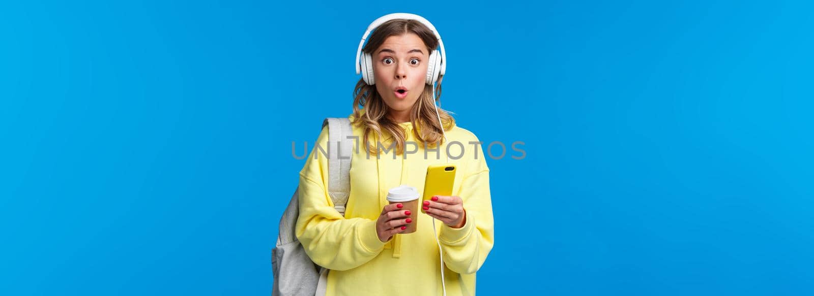 Surprised and amused young blond pretty girl look amazed camera say wow, wearing headphones and texting friend via smartphone, hold mobile phone, backpack and take-away coffee.