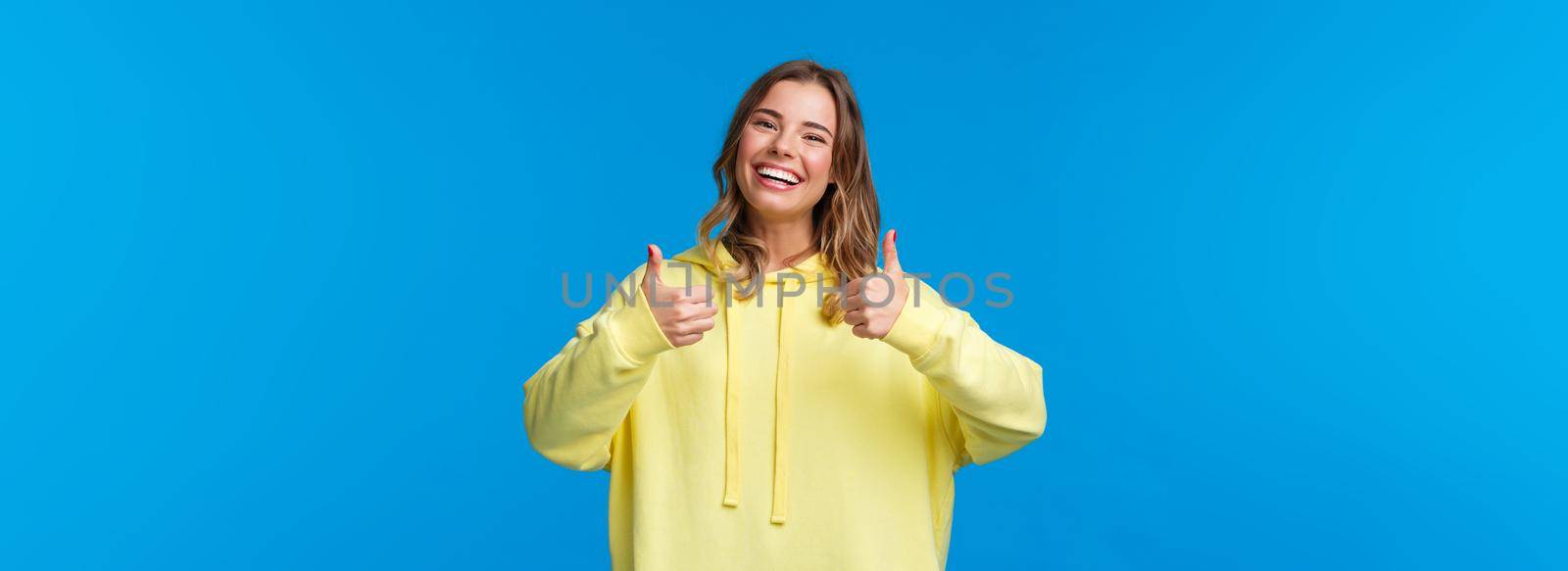 Sounds good Im in. Cheerful and optimistic good-looking young female blonde in yellow hoodie, show thumbs-up in approval, smiling nod acceptingly, like idea, blue background by Benzoix