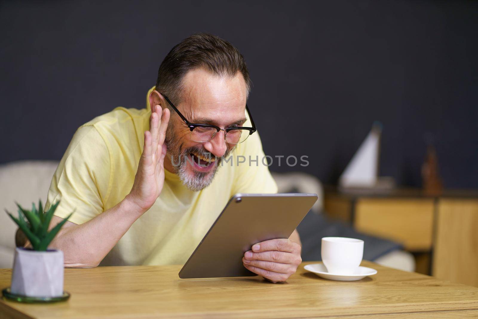 Happy middle aged man use digital tablet to communicate with his kids on vacation sitting at the home office desk drinking coffee wearing casual, glasses. Freelancer man work from home using gadget.