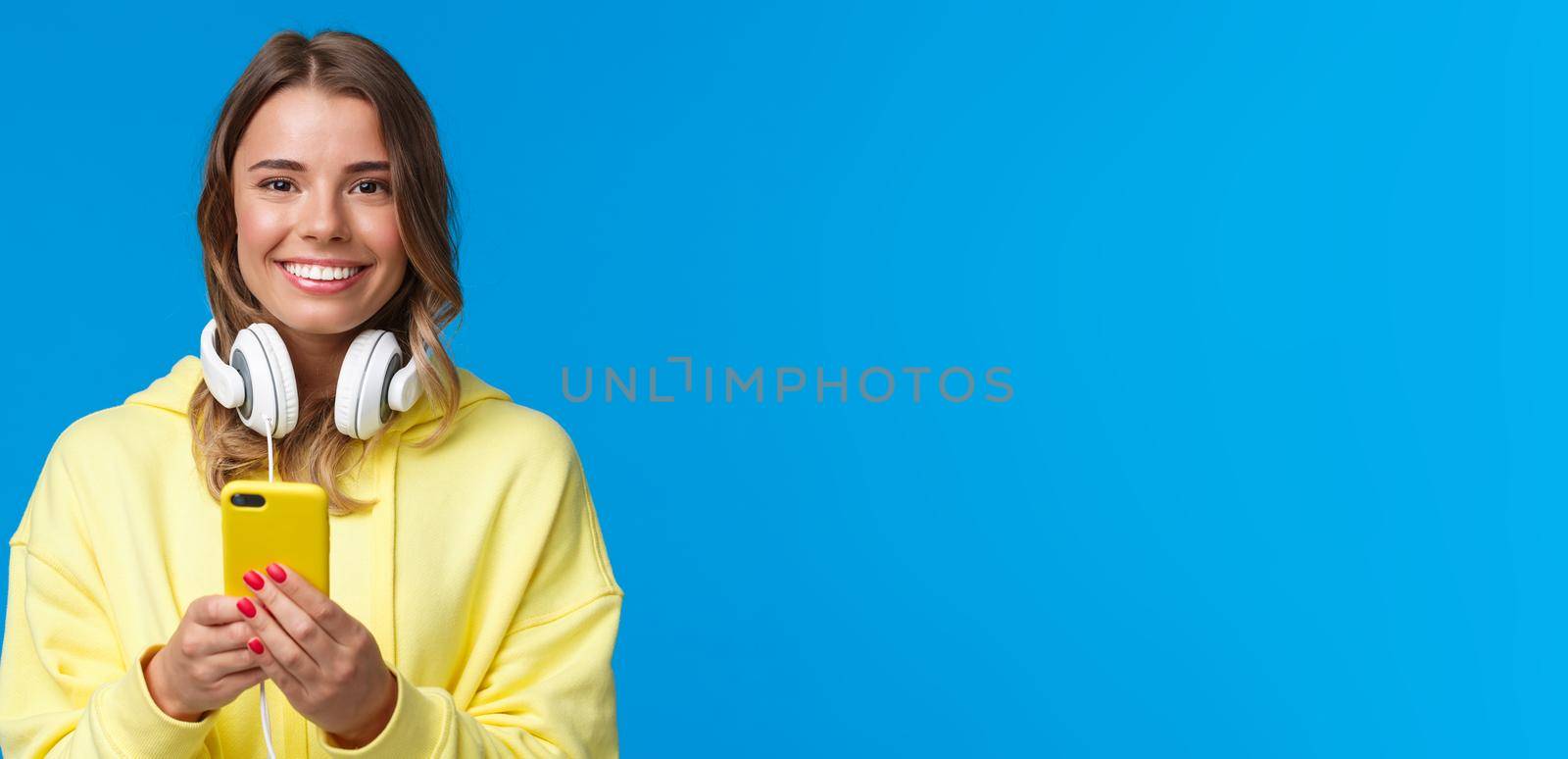 Close-up portrait of young blond european hipster girl with blond short hair and headphones over neck, holding smartphone, texting message, use mobile phone, blue background. Copy space