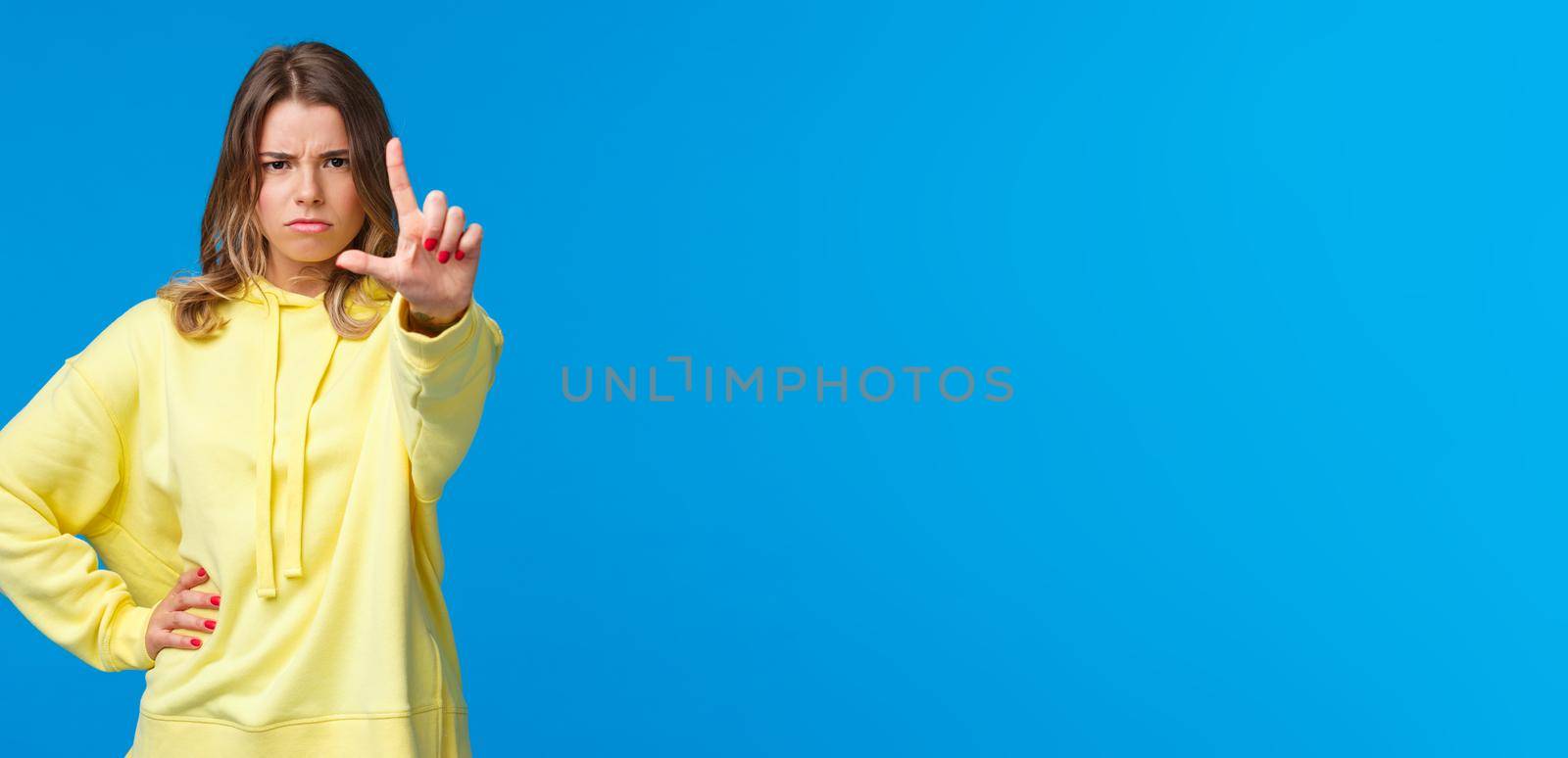 Stop right there. Serious-looking angry and bossy elder sister prohibit something, scolding kid as shaking finger with disapproval or disagreement, standing blue background.