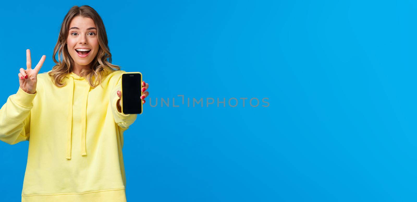 Optimistic cute blond european girl with short haircut, yellow hoodie, showing kawaii peace gesture and mobile phone display as using photo filter to edit and post pic online, blue background.