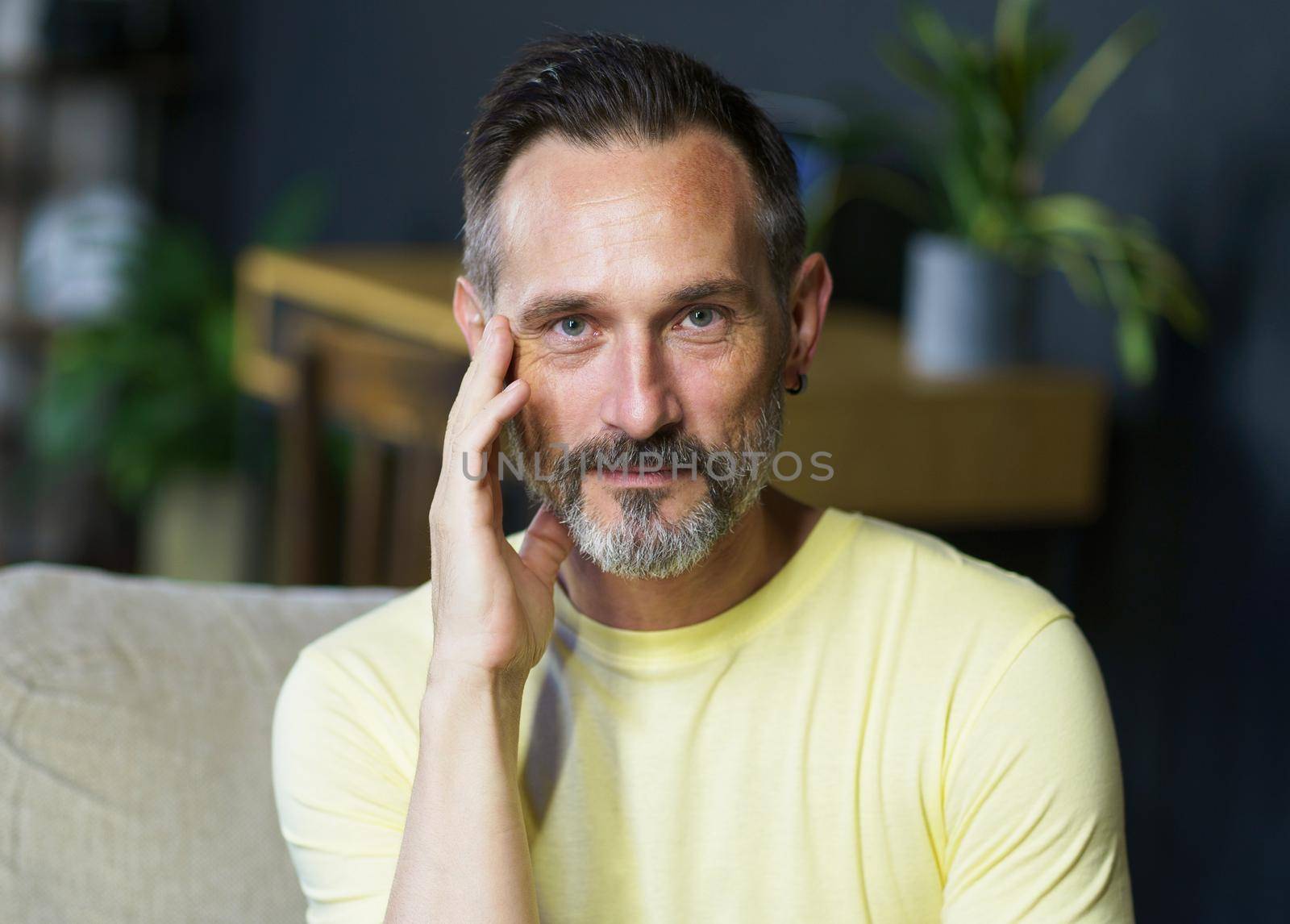 Relaxed handsome middle aged grey haired man sitting on the sofa touching his face with a hand looking at camera wearing casual. Thoughtful freelancer mature man working from home.