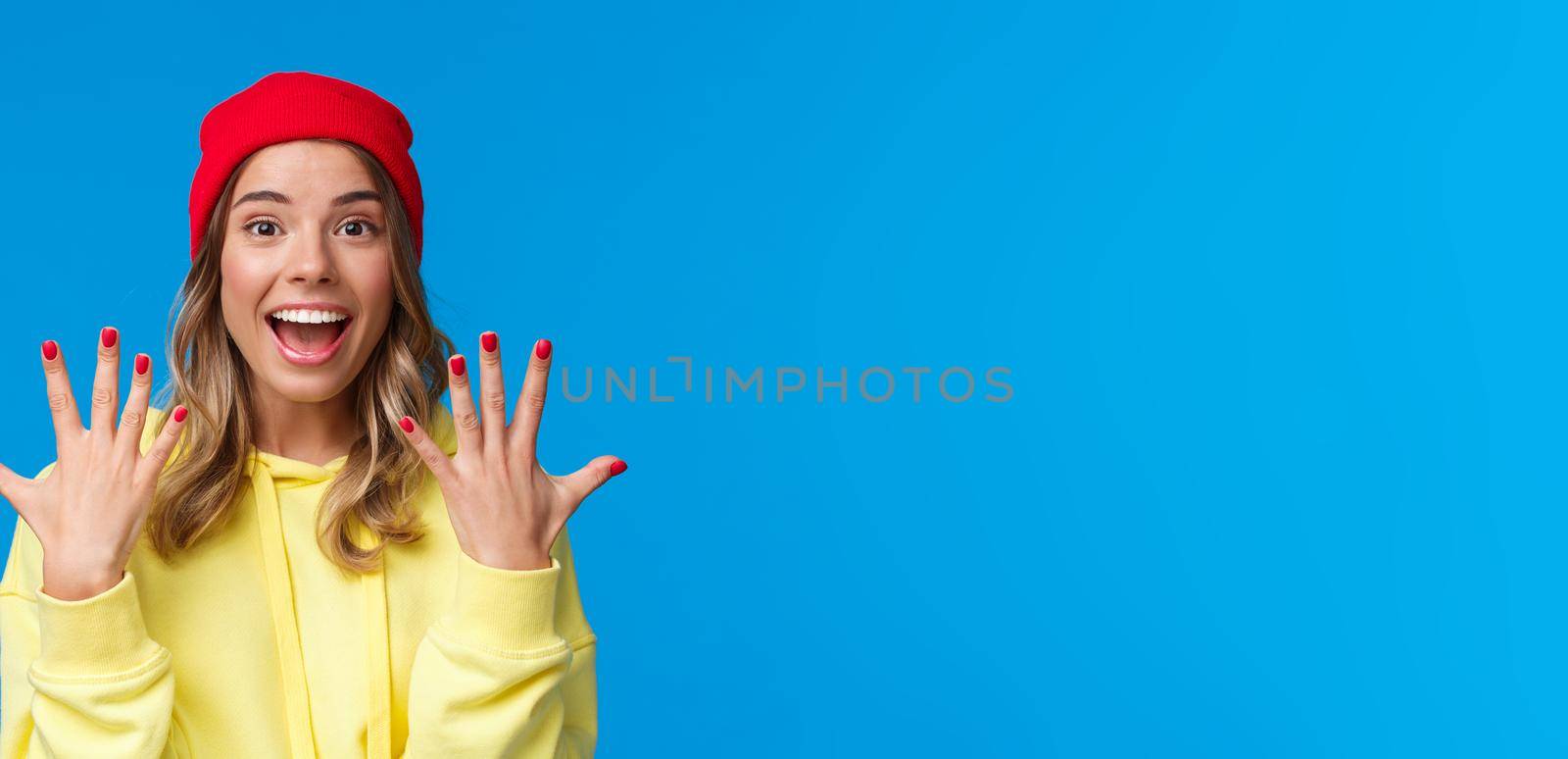 Portrait of amused good-looking blonde hipster girl smiling joyfully, explain something showing number ten, tenth with hands and look camera upbeat, order dozen, stand blue background.