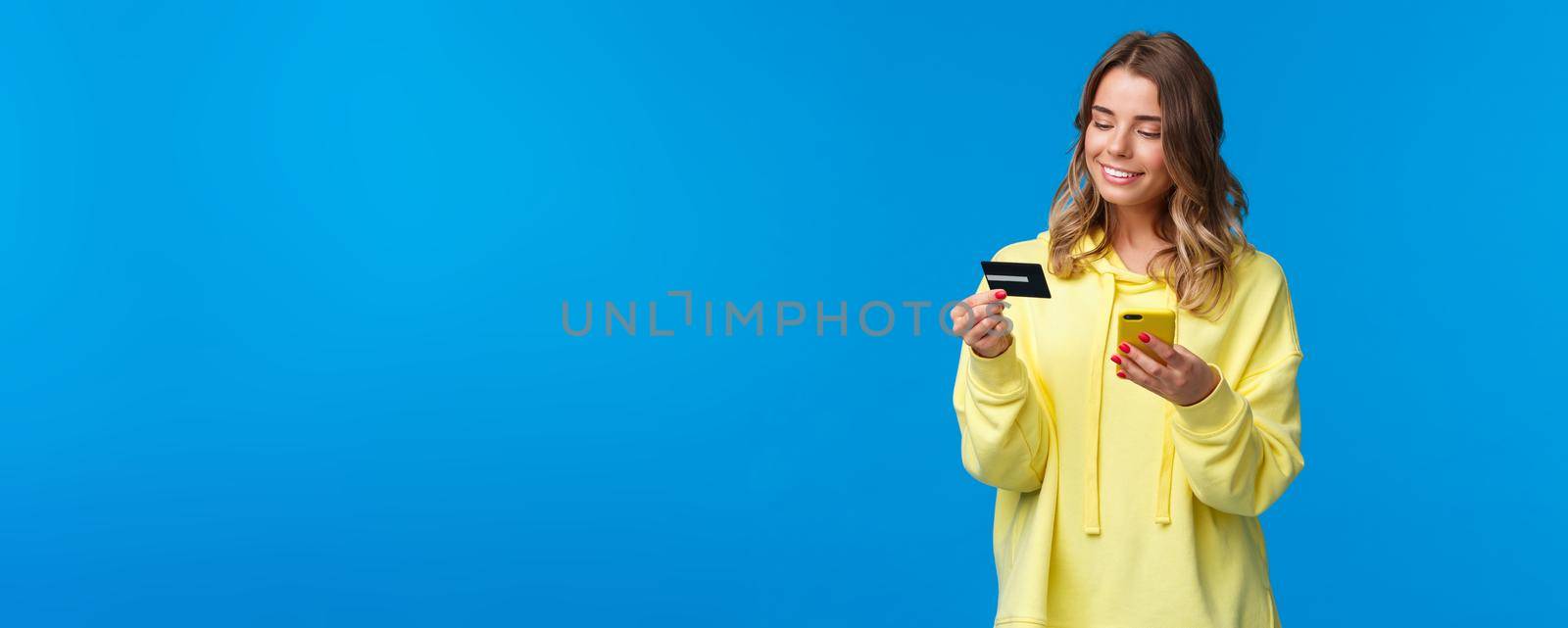 Young caucasian female with blond short hair using mobile phone and insert digit numbers of credit card to pay for her online purchase, shopping in internet store, smiling pleased by Benzoix