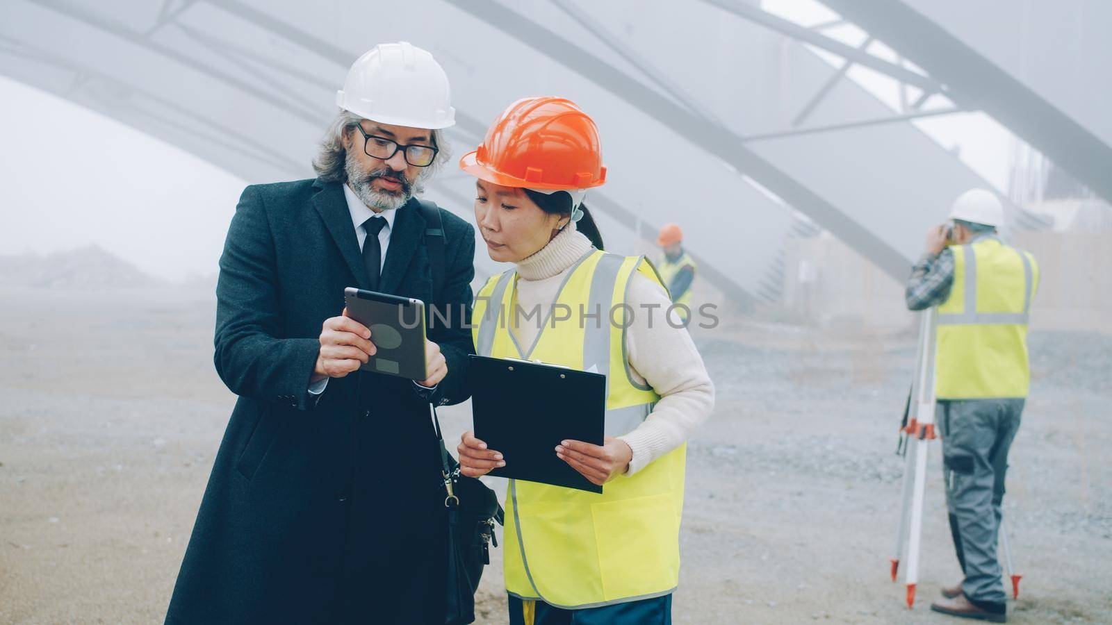 Asian woman engineer talking to Caucasian man investor discussing construction project reading papers and using tablet by silverkblack
