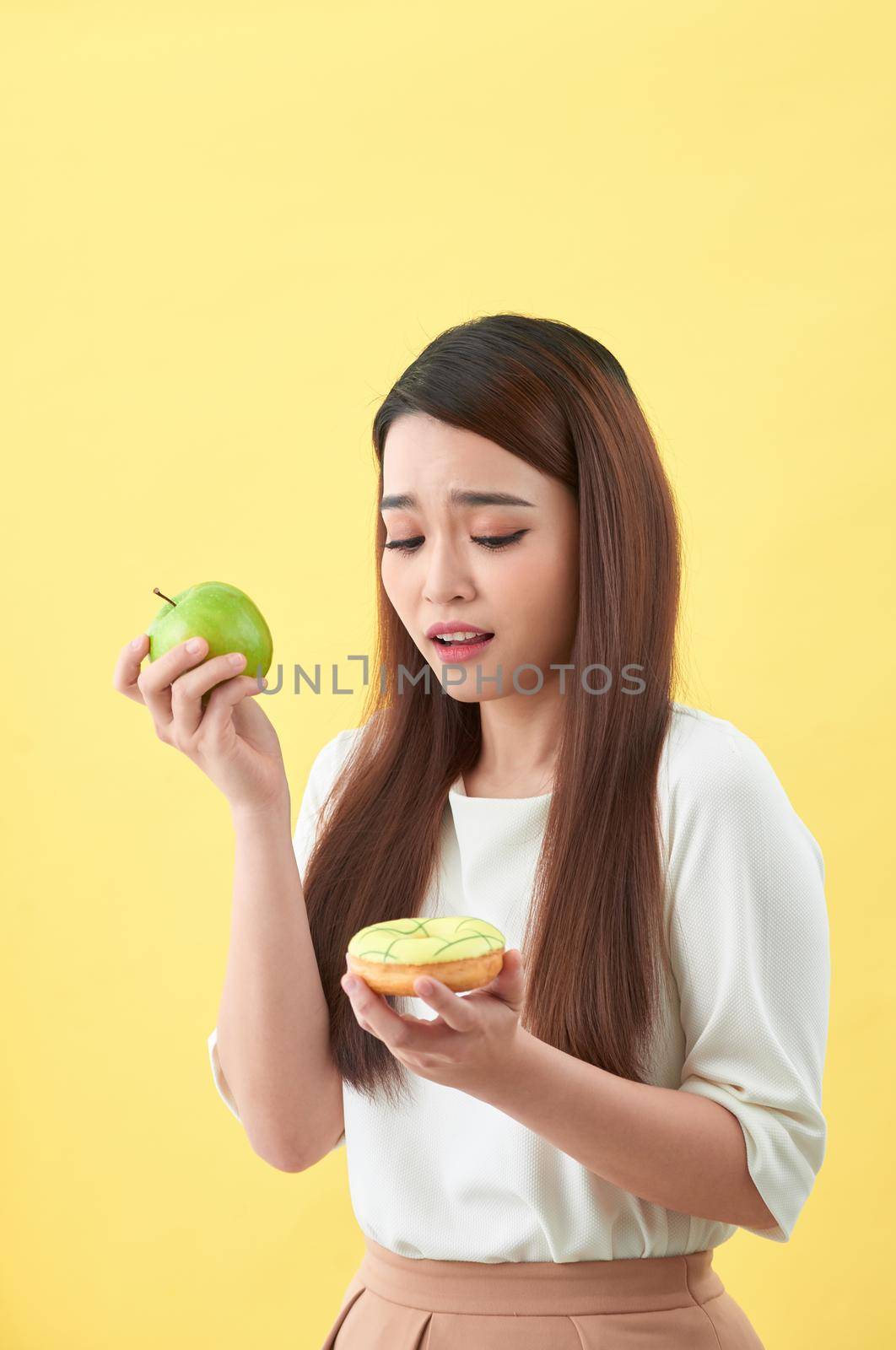 Portrait of a smiling young asian woman choosing between donut and green apple isolated over yellow background