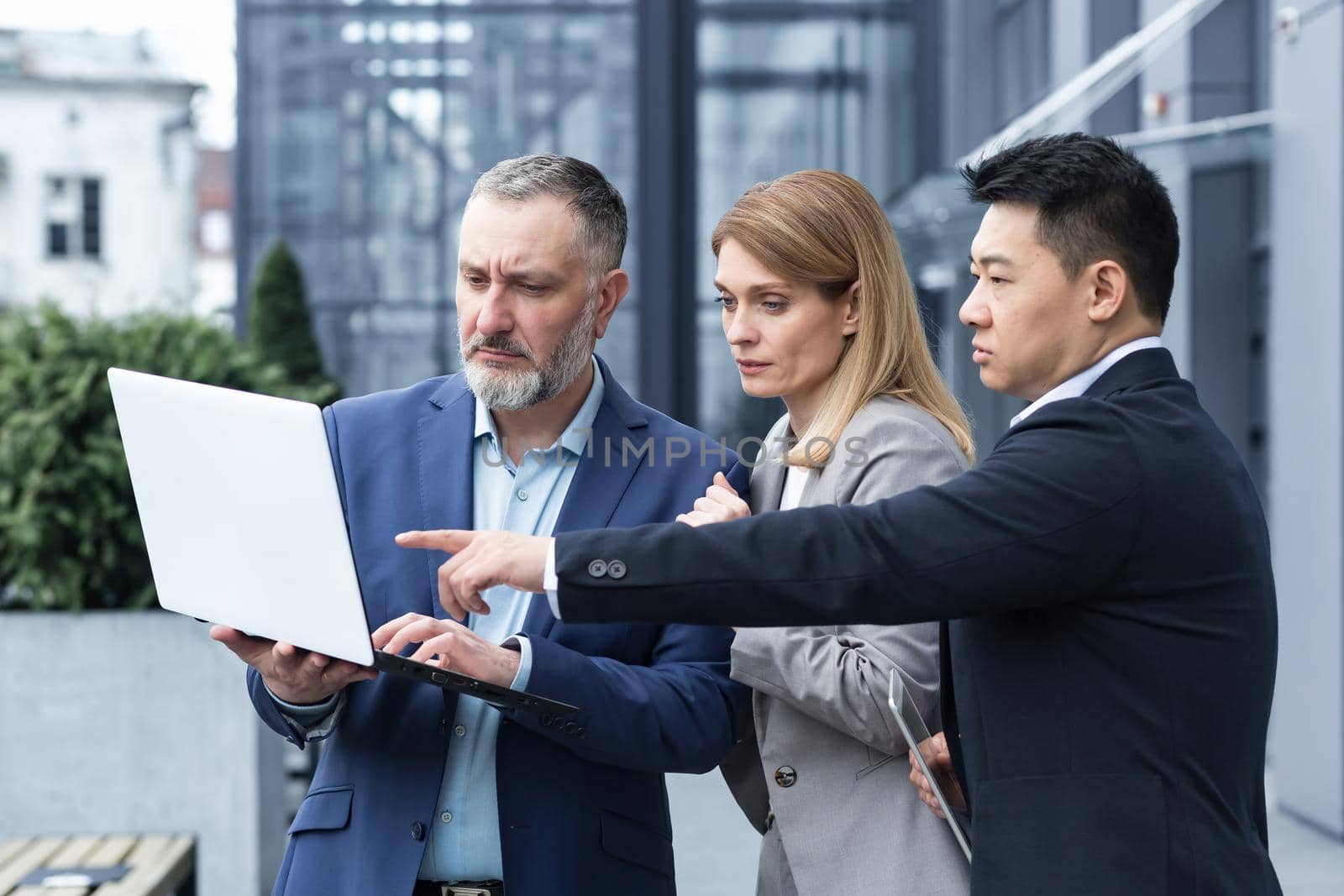 Successful business team, three colleagues businessman and businesswoman outside office building discussing current plans and management, looking at laptop screen, discussing and consulting