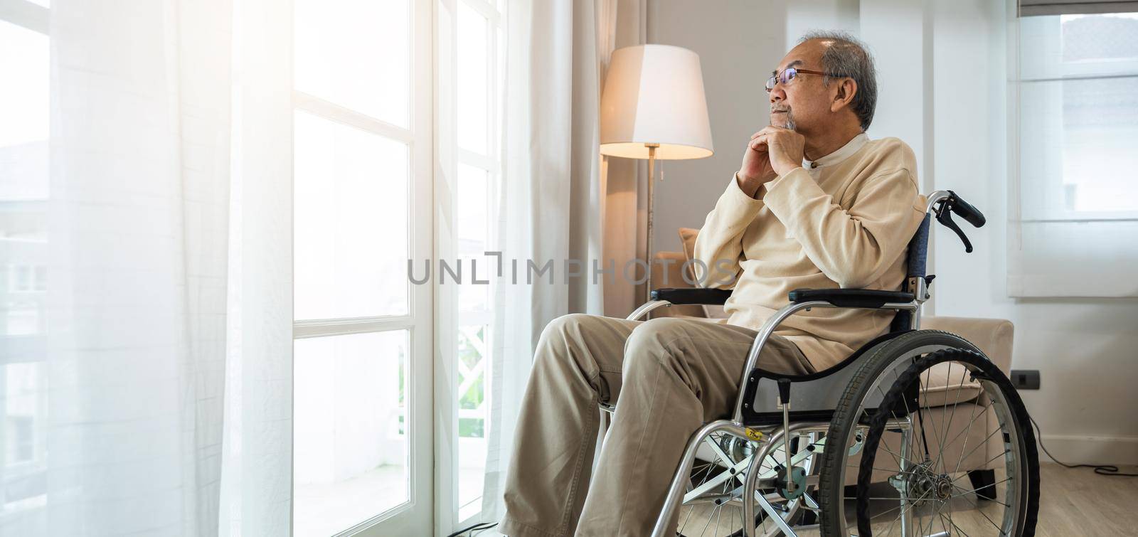 Asian senior man disabled sitting alone in wheelchair looking through window at hospital by Sorapop