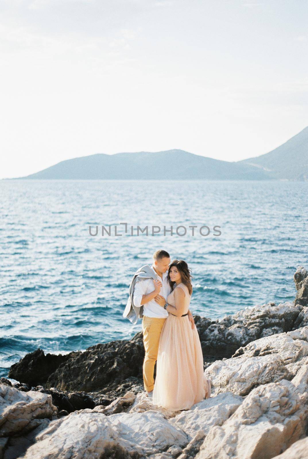 Groom with a jacket on his shoulder hugs bride on a rocky seashore. High quality photo