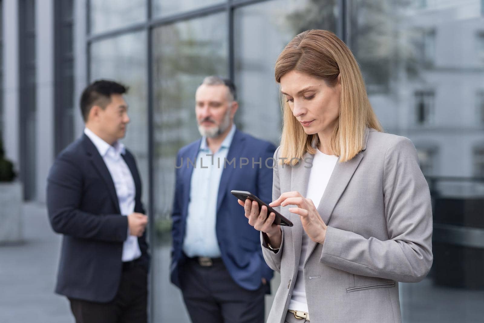 A successful and experienced business woman outside the office building is reading from a tablet computer, an employee with colleagues in business suits on a break