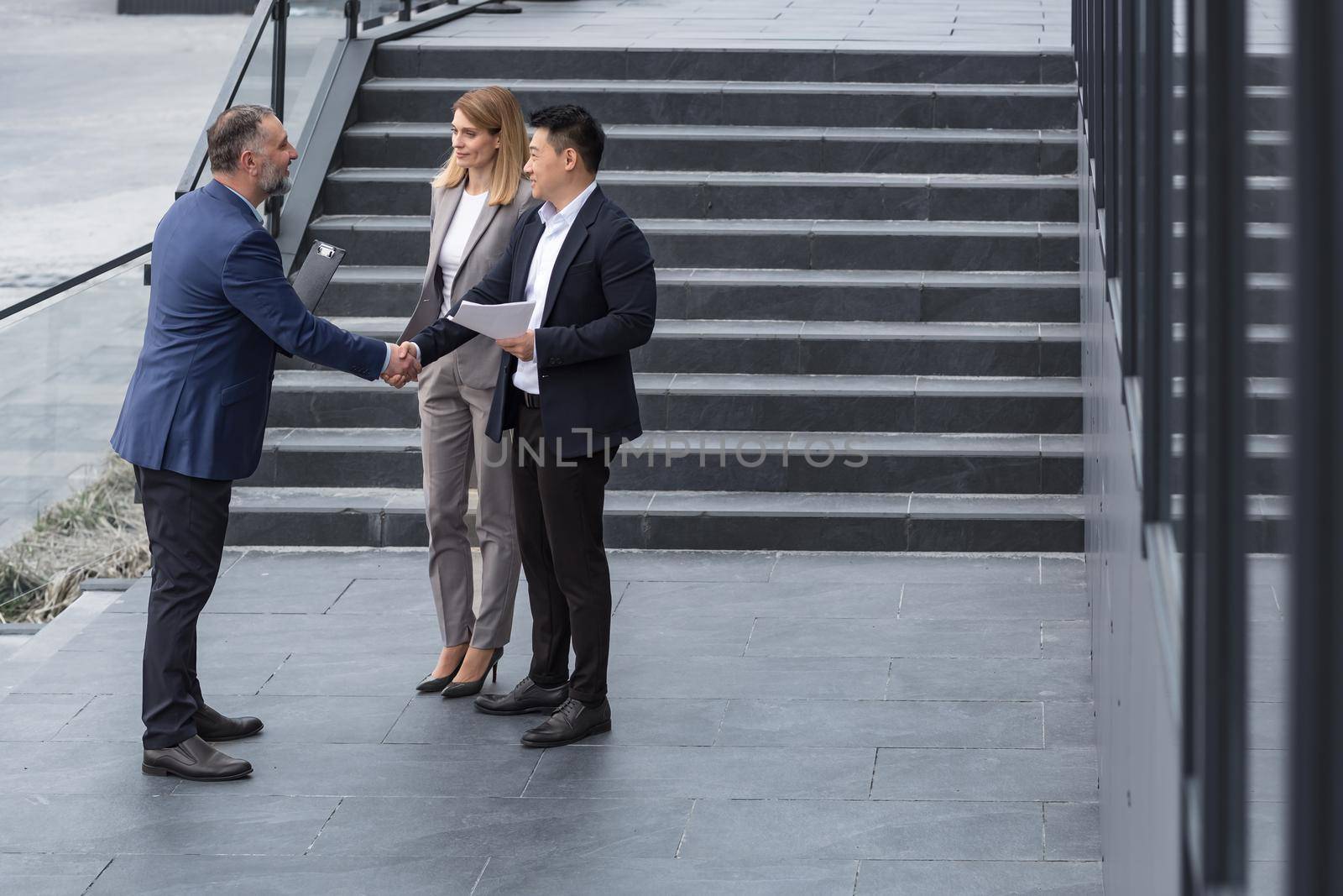 Meeting of three colleagues from outside the office building, experienced and mature IT specialists, greeting and shaking hands, business persons in business suits, diverse group of people