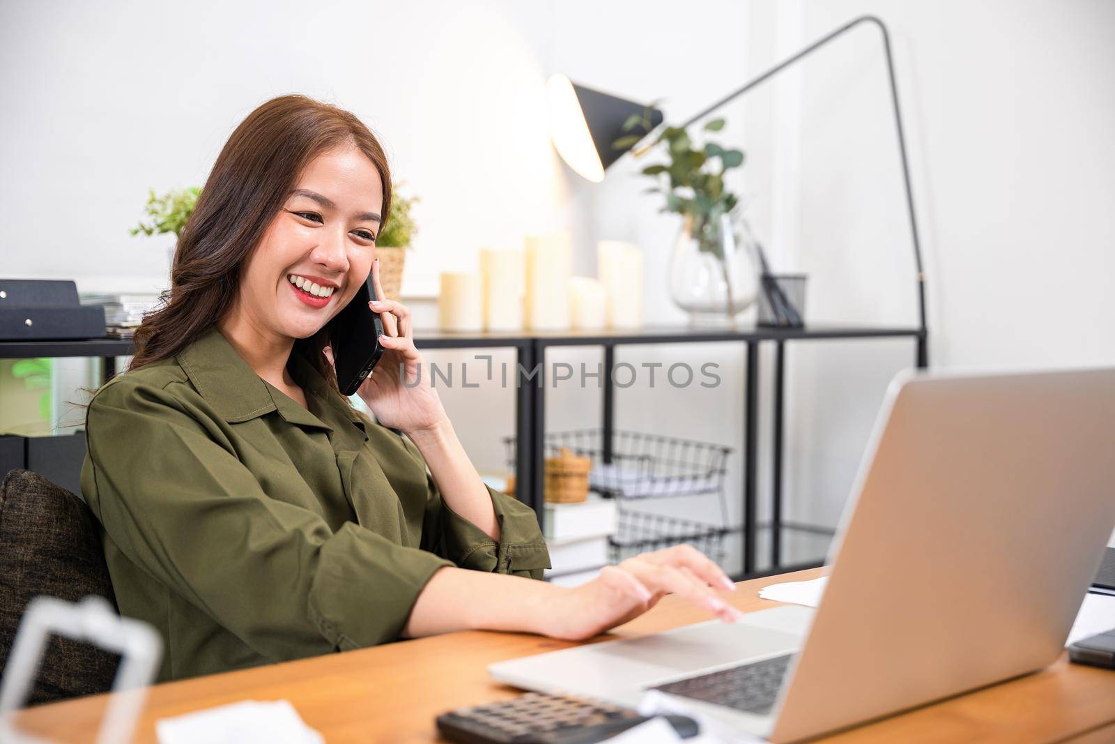 woman working on laptop computer at home office while talking on mobile phone by Sorapop