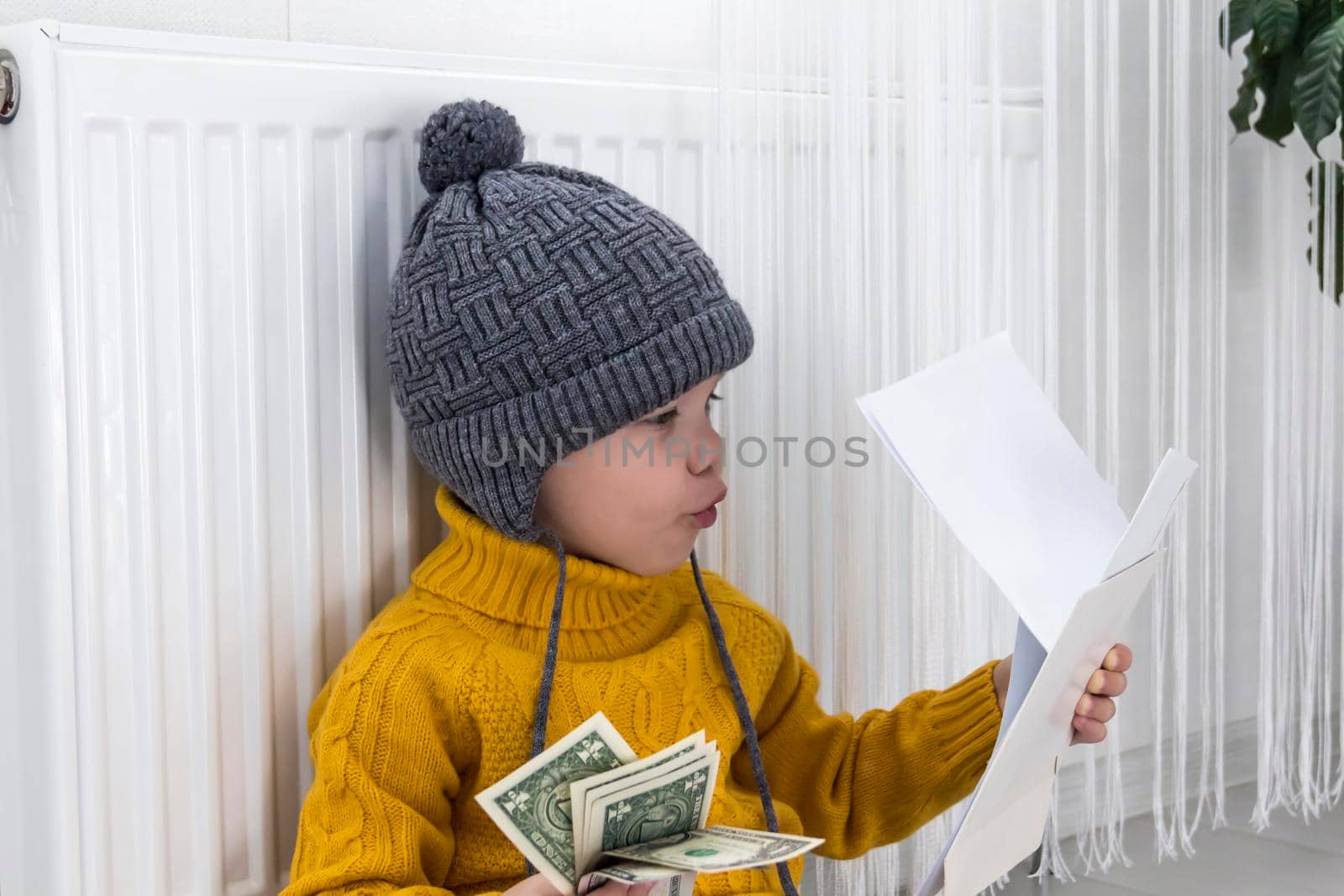 A little boy in a yellow sweater and hat is counting money and studying heating bills, near a heater with a thermostat..