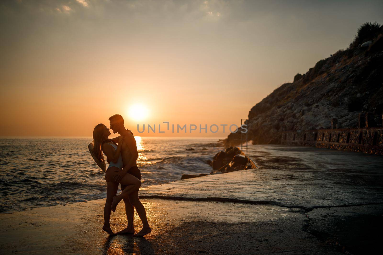 Beautiful couple in love on the background of the sunset by the sea. Young woman and man hugging by the sea at sunset