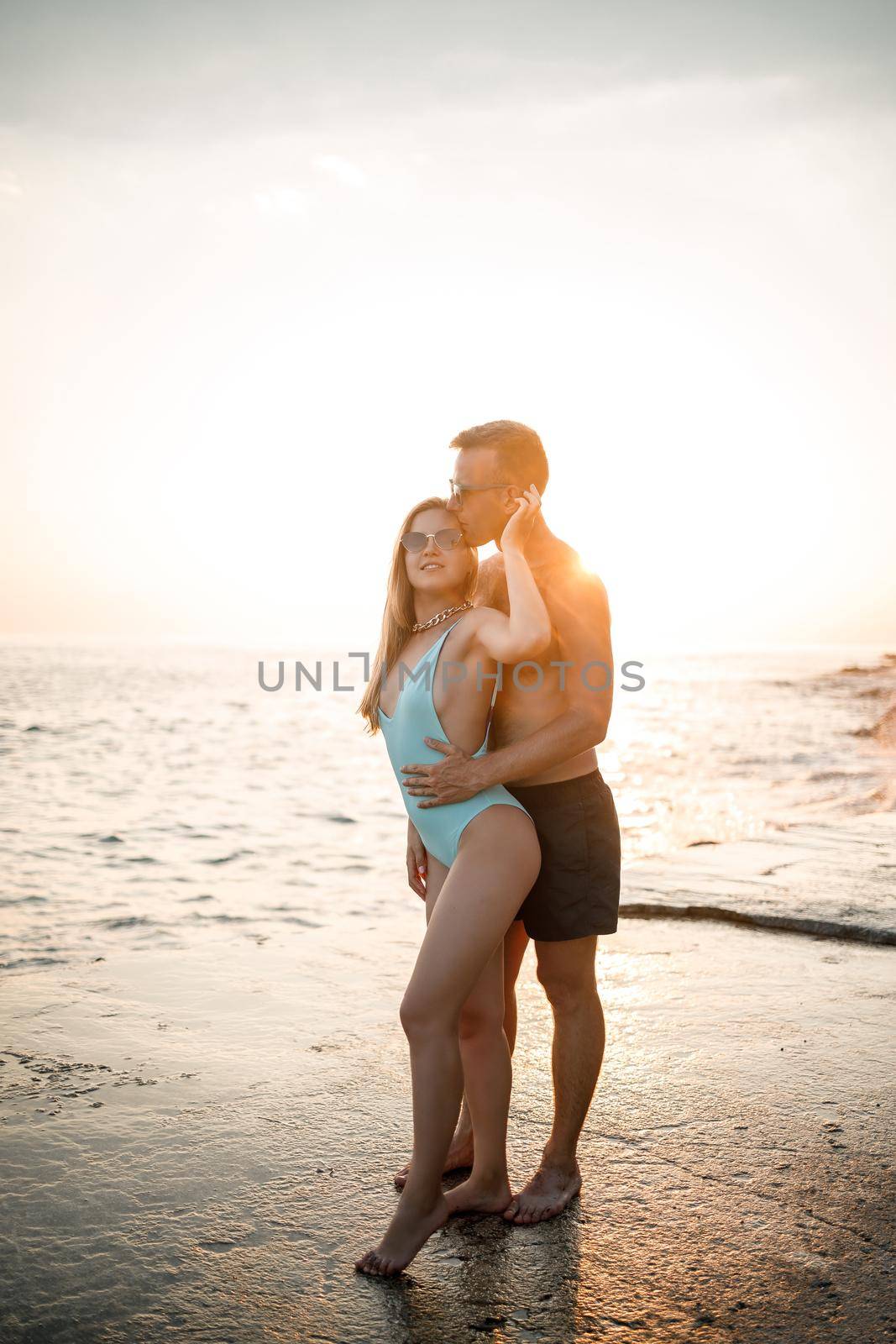 A loving couple walks along the beach by the sea. Young family at sunset by the Mediterranean Sea. Vacation concept. A woman in a swimsuit and a man in shorts at sunset by the sea. Selective focus.