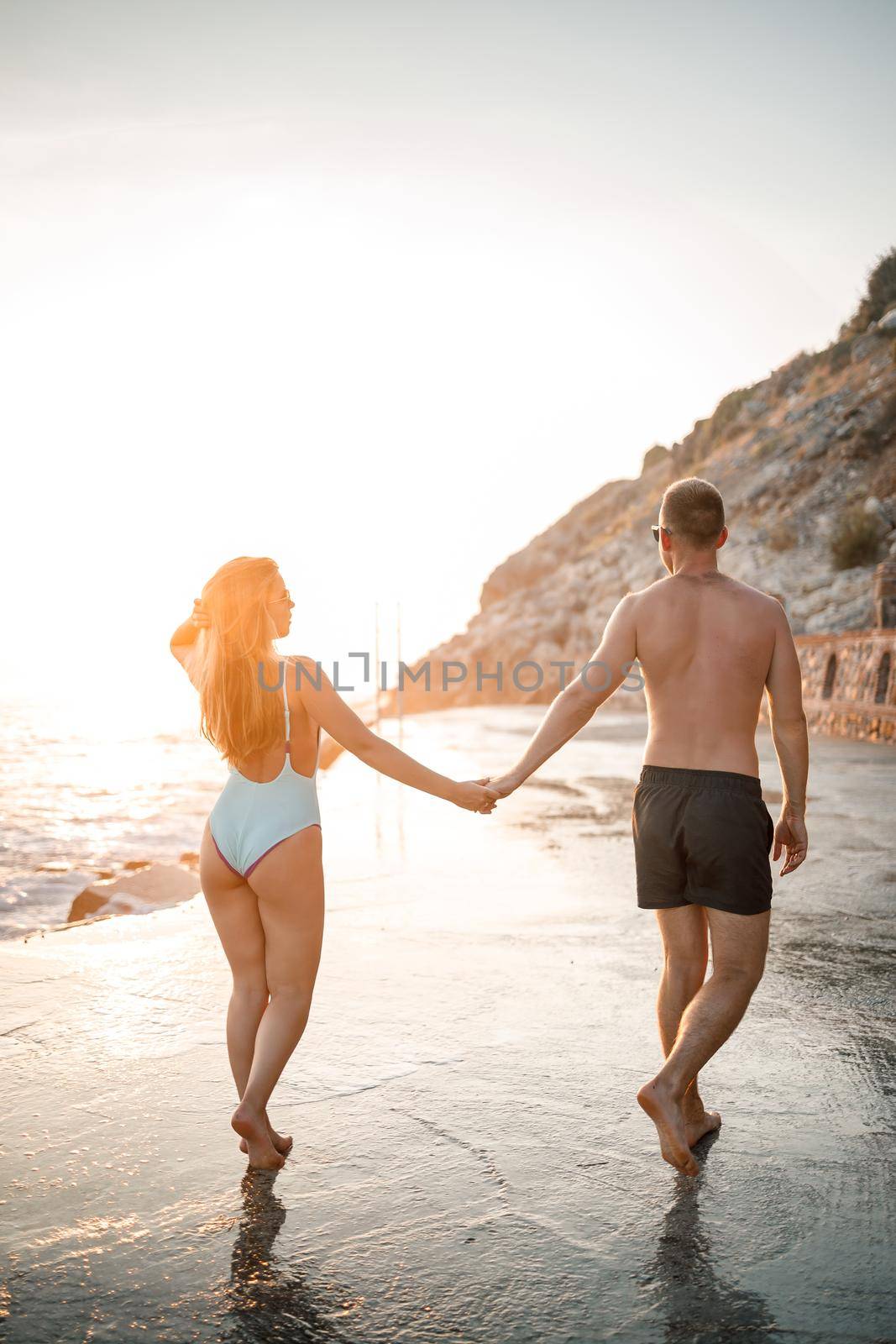 A loving couple walks along the beach by the sea. Young family at sunset by the Mediterranean Sea. Vacation concept. A woman in a swimsuit and a man in shorts at sunset by the sea. Selective focus.