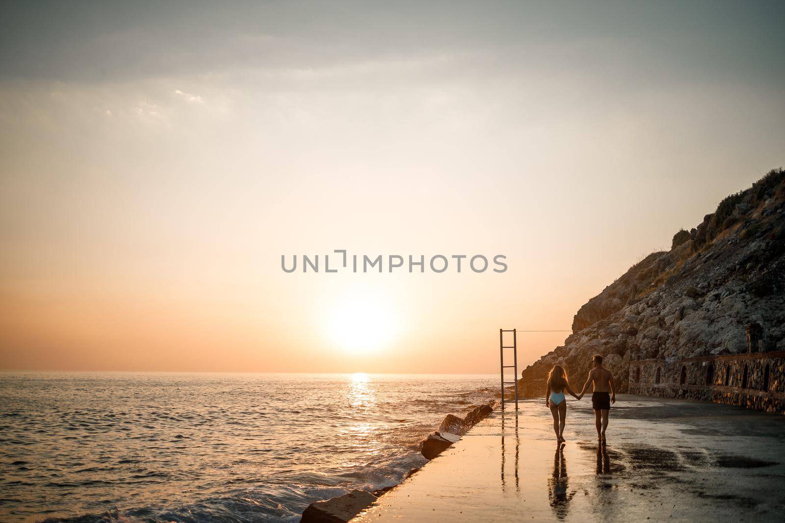 A loving couple walks along the beach by the sea. Young family at sunset by the Mediterranean Sea. Vacation concept. A woman in a swimsuit and a man in shorts at sunset by the sea. Selective focus.