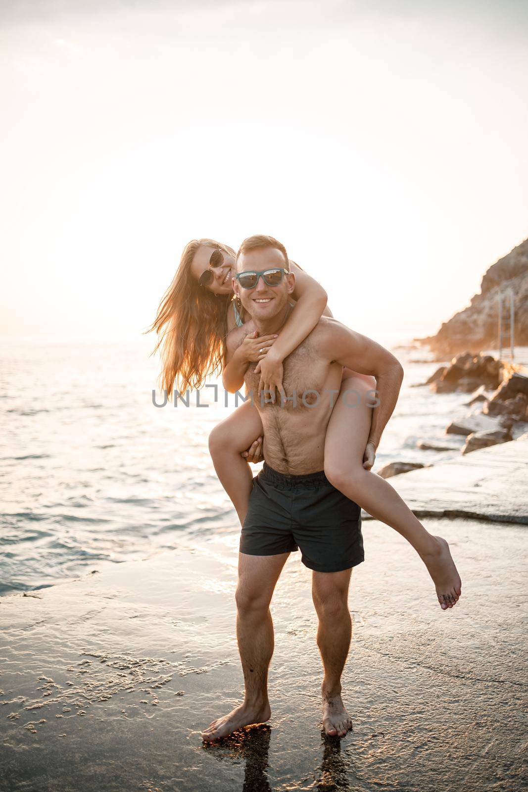 A loving couple walks along the beach by the sea. Young family at sunset by the Mediterranean Sea. Vacation concept. A woman in a swimsuit and a man in shorts at sunset by the sea. Selective focus. by Dmitrytph
