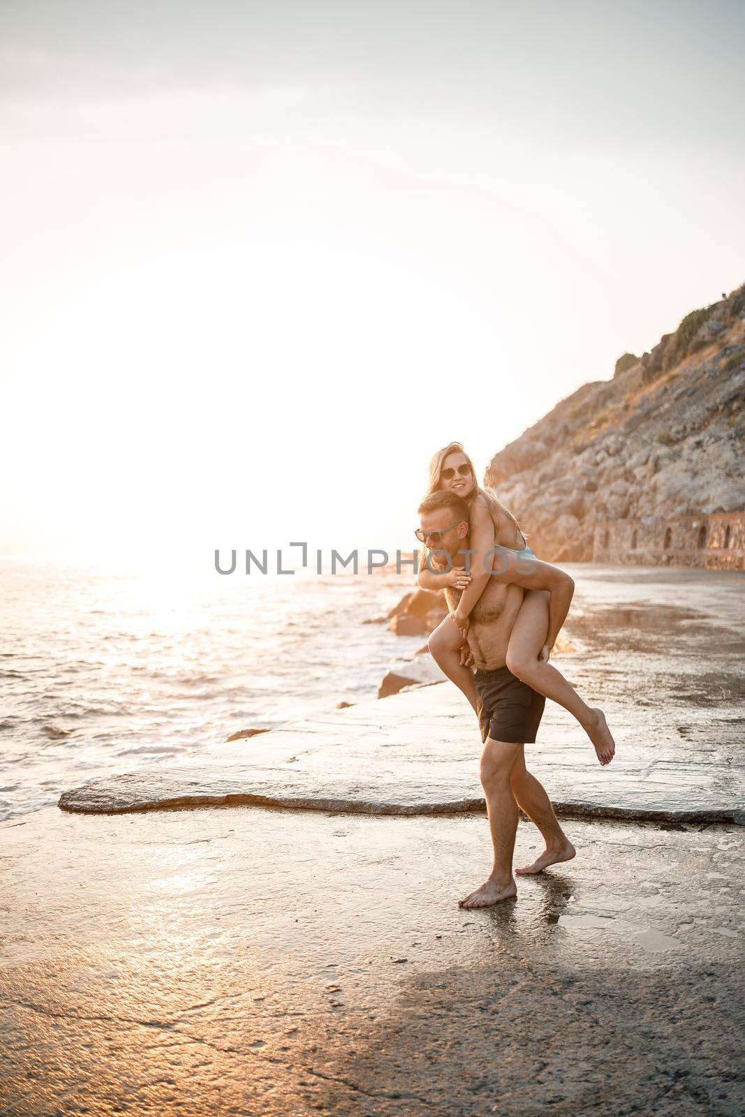 A loving couple walks along the beach by the sea. Young family at sunset by the Mediterranean Sea. Vacation concept. A woman in a swimsuit and a man in shorts at sunset by the sea. Selective focus. by Dmitrytph