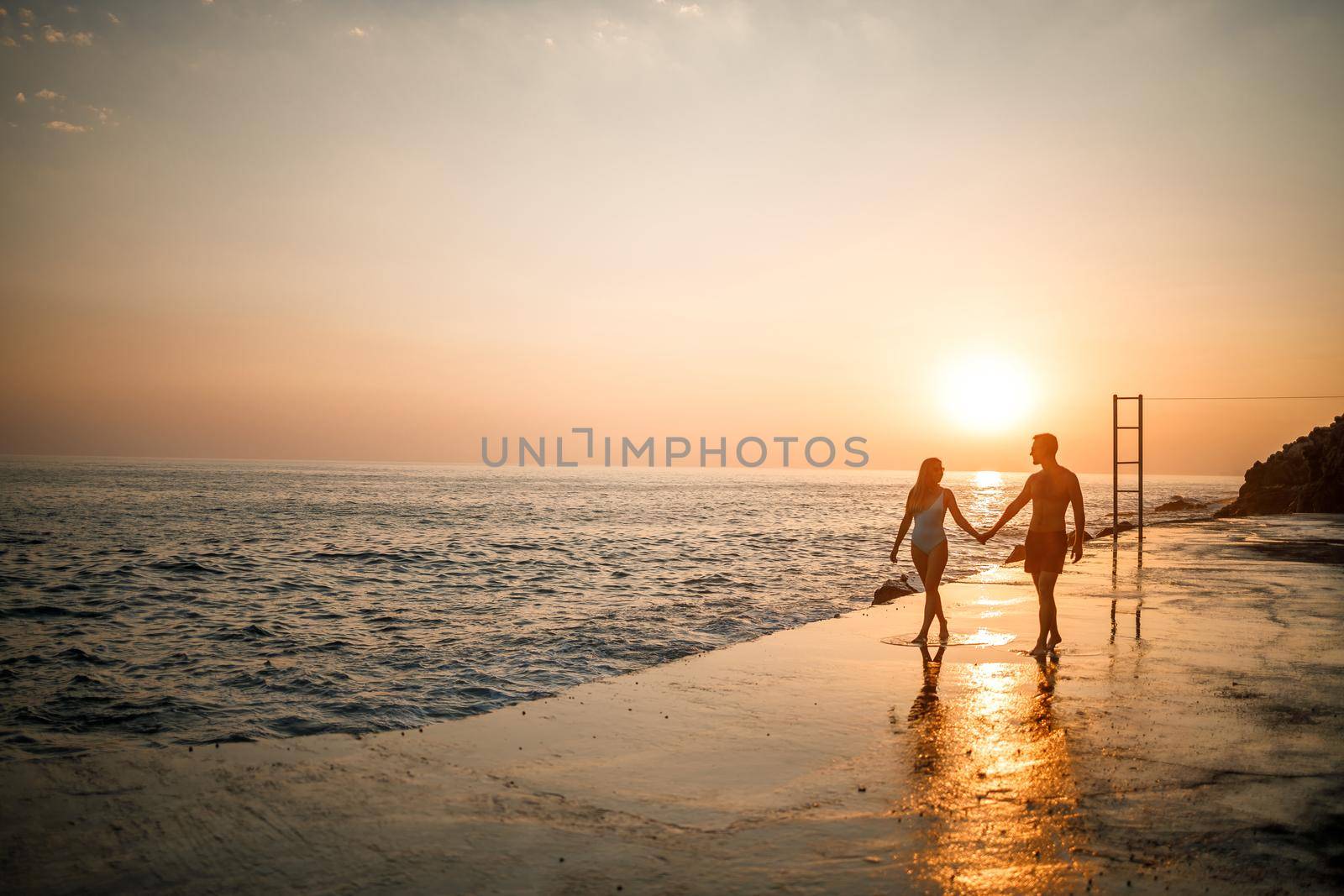 A loving couple walks along the beach by the sea. Young family at sunset by the Mediterranean Sea. Vacation concept. A woman in a swimsuit and a man in shorts at sunset by the sea. Selective focus.