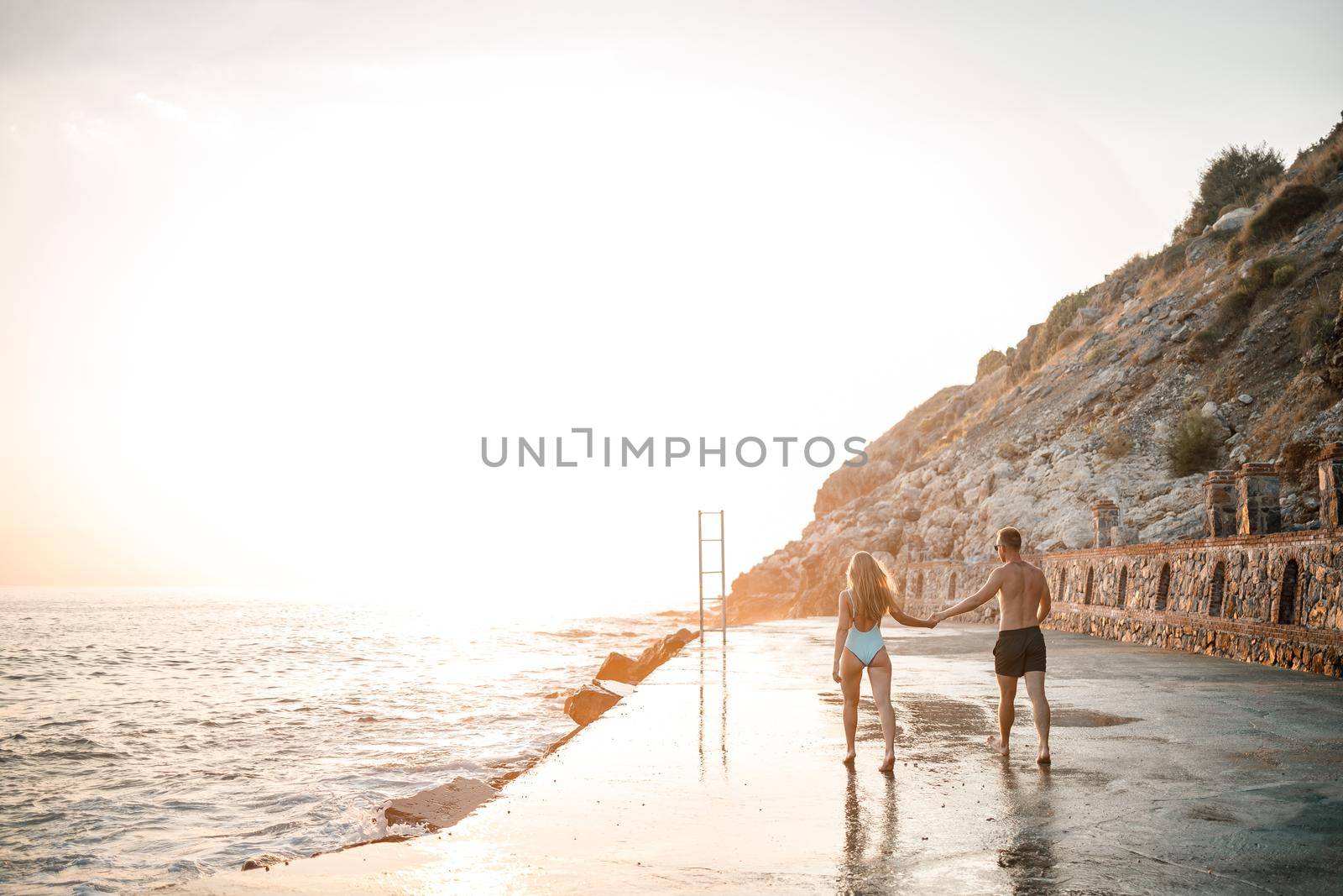 A loving couple walks along the beach by the sea. Young family at sunset by the Mediterranean Sea. Vacation concept. A woman in a swimsuit and a man in shorts at sunset by the sea. Selective focus.