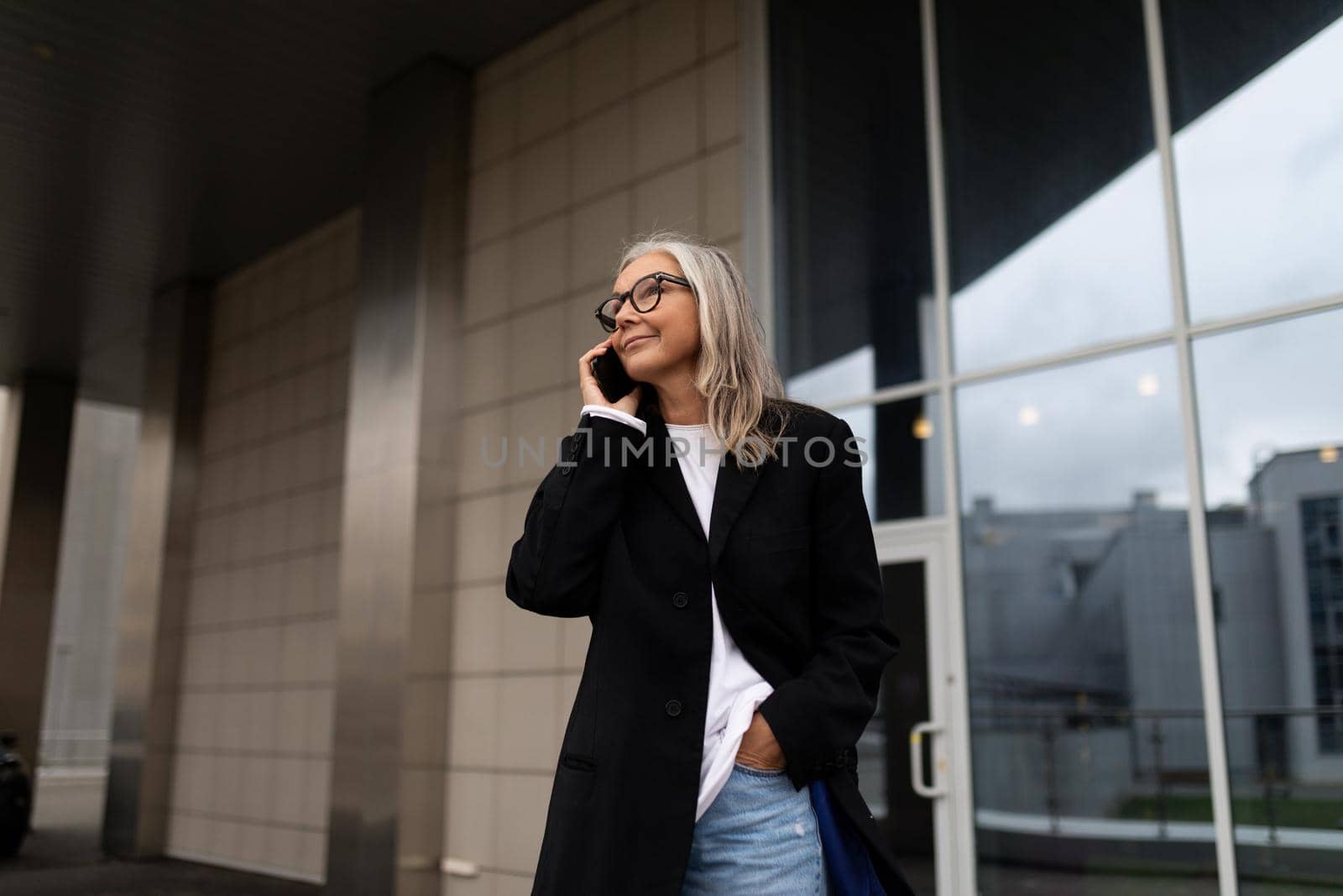 successful elderly businesswoman talking on a mobile phone against the backdrop of the facade of the office center by TRMK
