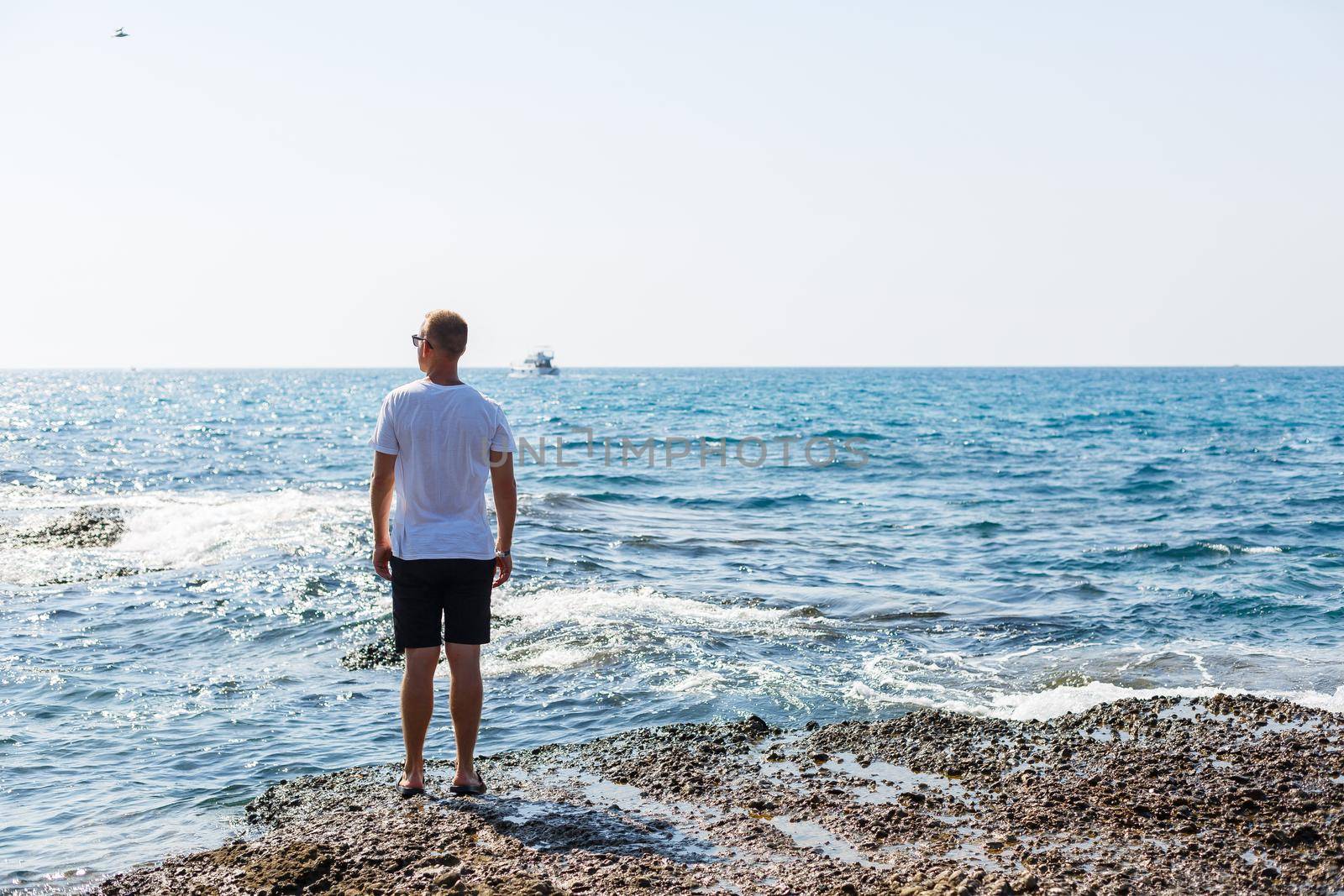 Young attractive man in sunglasses in a white t-shirt and shorts stands on the shore of the mediterranean sea by Dmitrytph