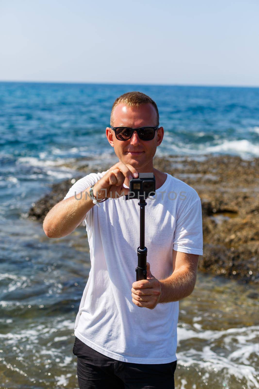 A man in a white T-shirt holds an action camera in his hands and shoots a beautiful view of the sea by Dmitrytph