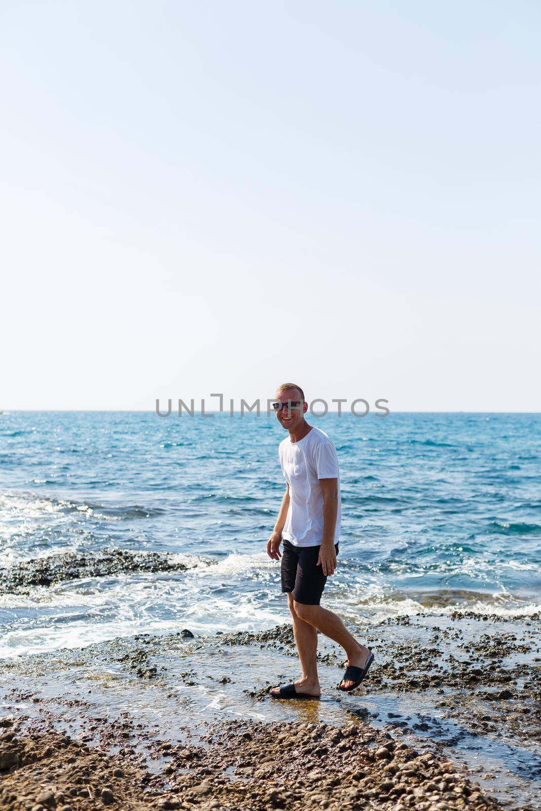 Young attractive man in sunglasses in a white t-shirt and shorts stands on the shore of the mediterranean sea by Dmitrytph