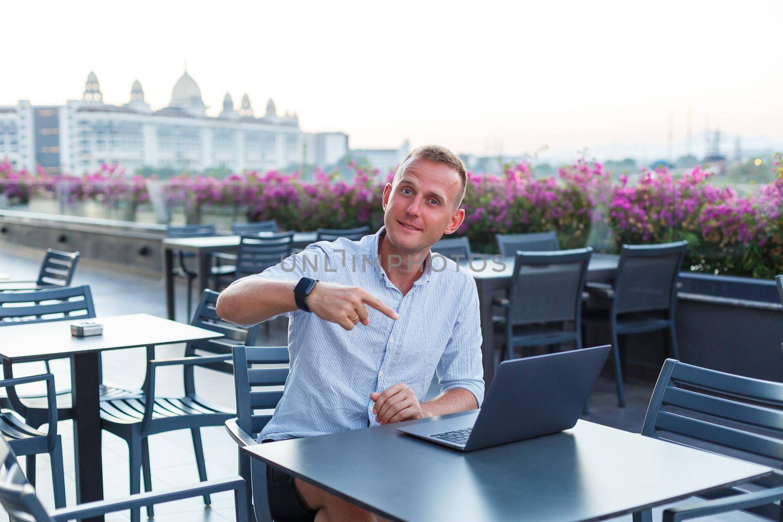 Successful young executive looking while sitting at a table with a laptop on a hotel terrace and finding time for remote control of employees while on vacation in an exotic resort