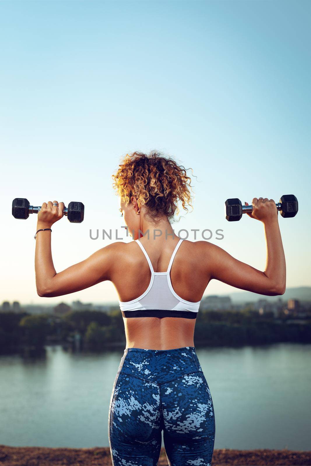 Rear view of a young fitness woman doing workout with dumbbells by the river in a sunset.