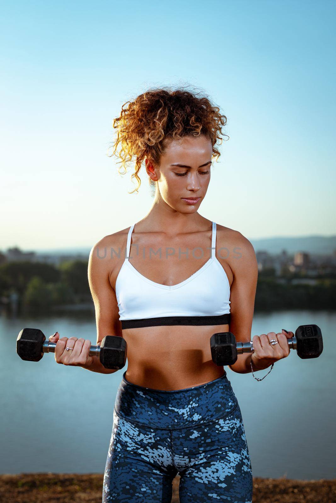 Young fitness woman doing workout with dumbbells by the river in a sunset.