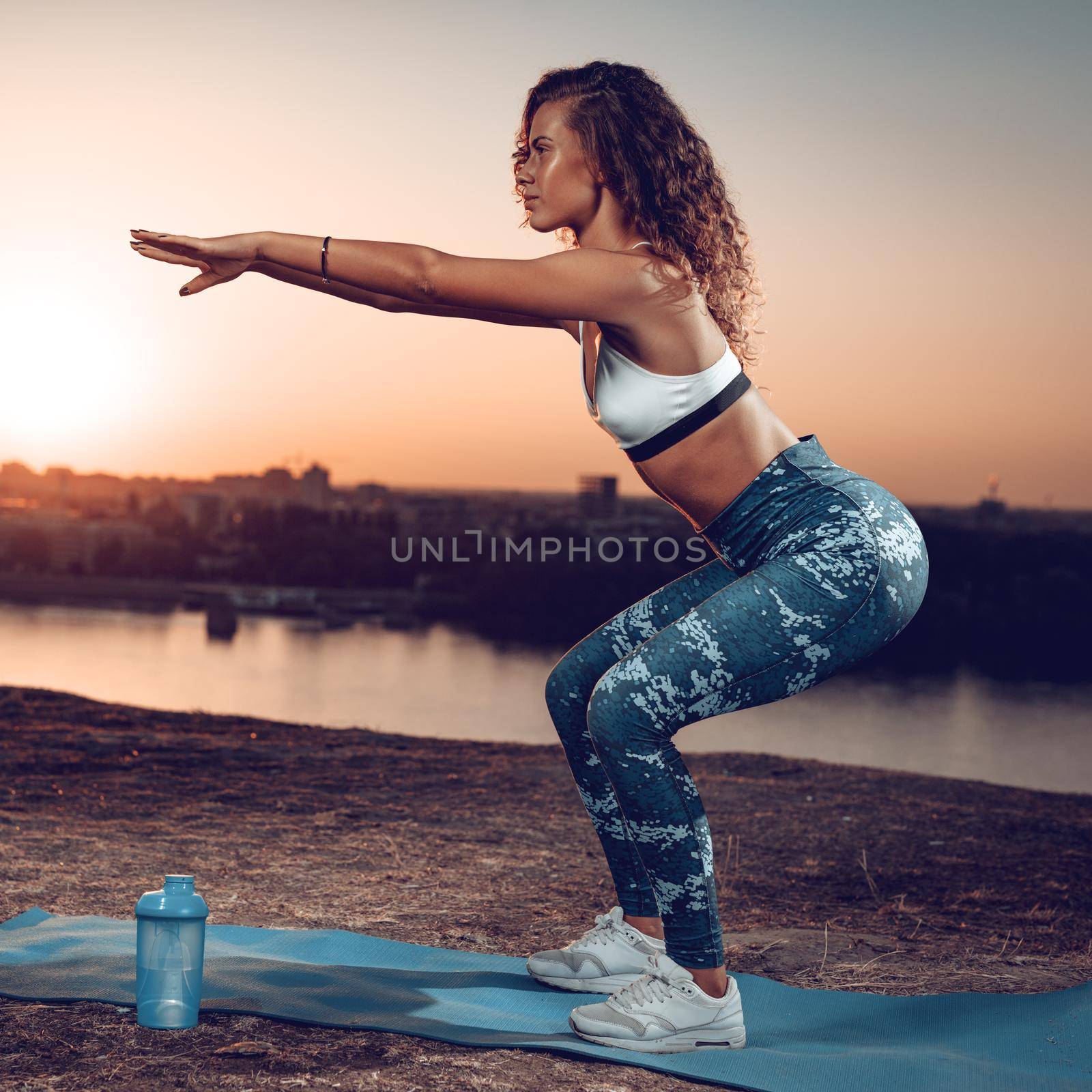 A profile of a young fitness woman doing exercises by the river in a sunset.