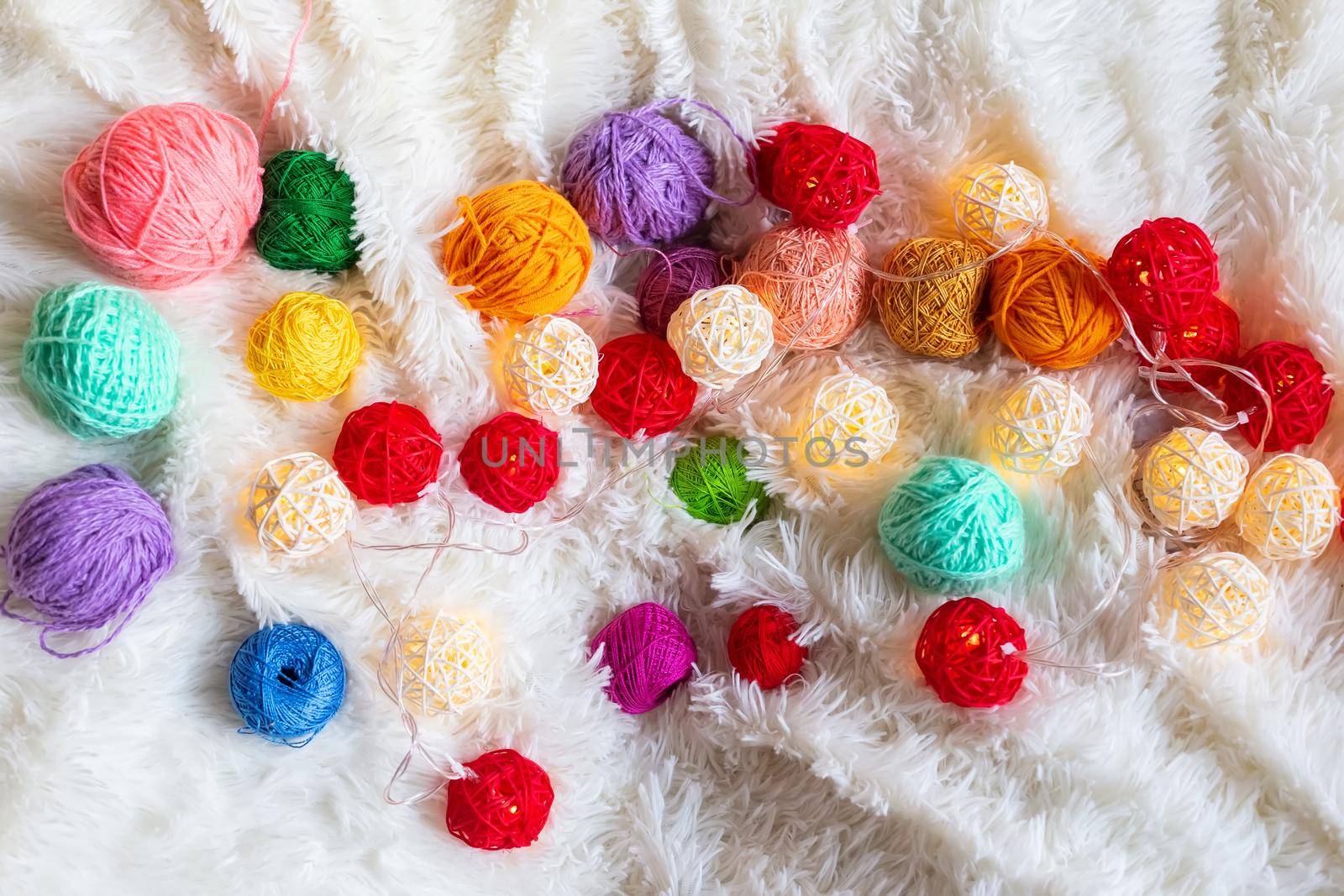 A lot of colorful knitting yarn and a luminous garland in the form of balls lie on a white fluffy blanket. Cozy still life made of yarn. Top view. Copy space