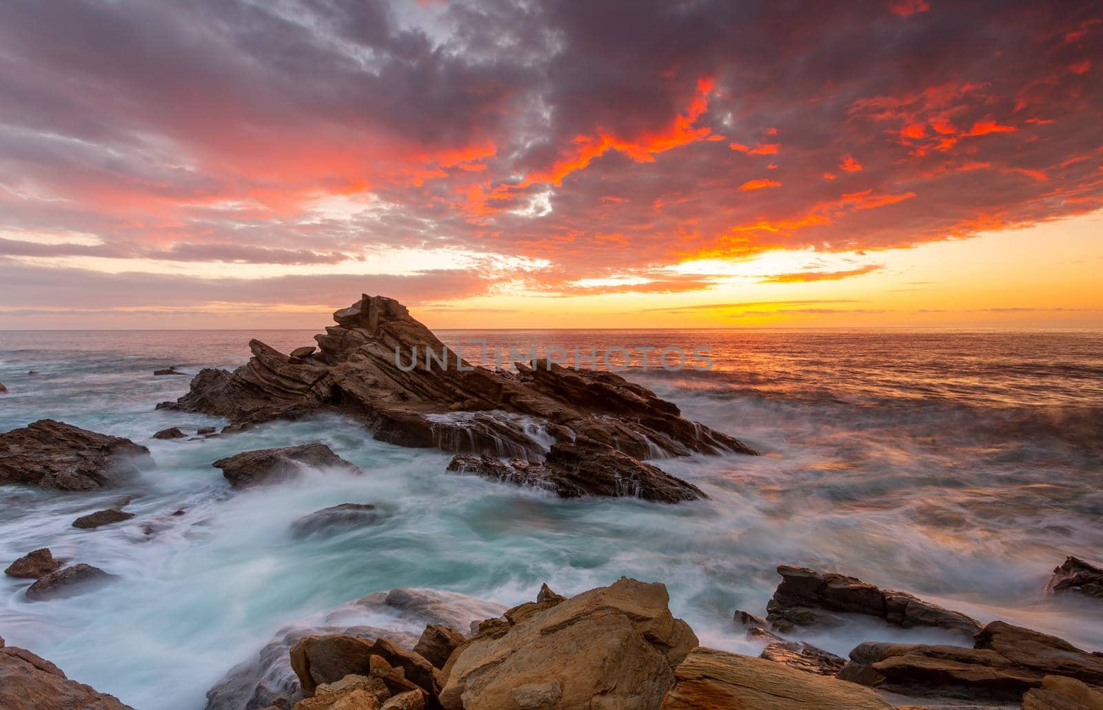 Sunrise over the ocean with rocky beach coast and flowing waves moving around jagged rocks in the foreground.