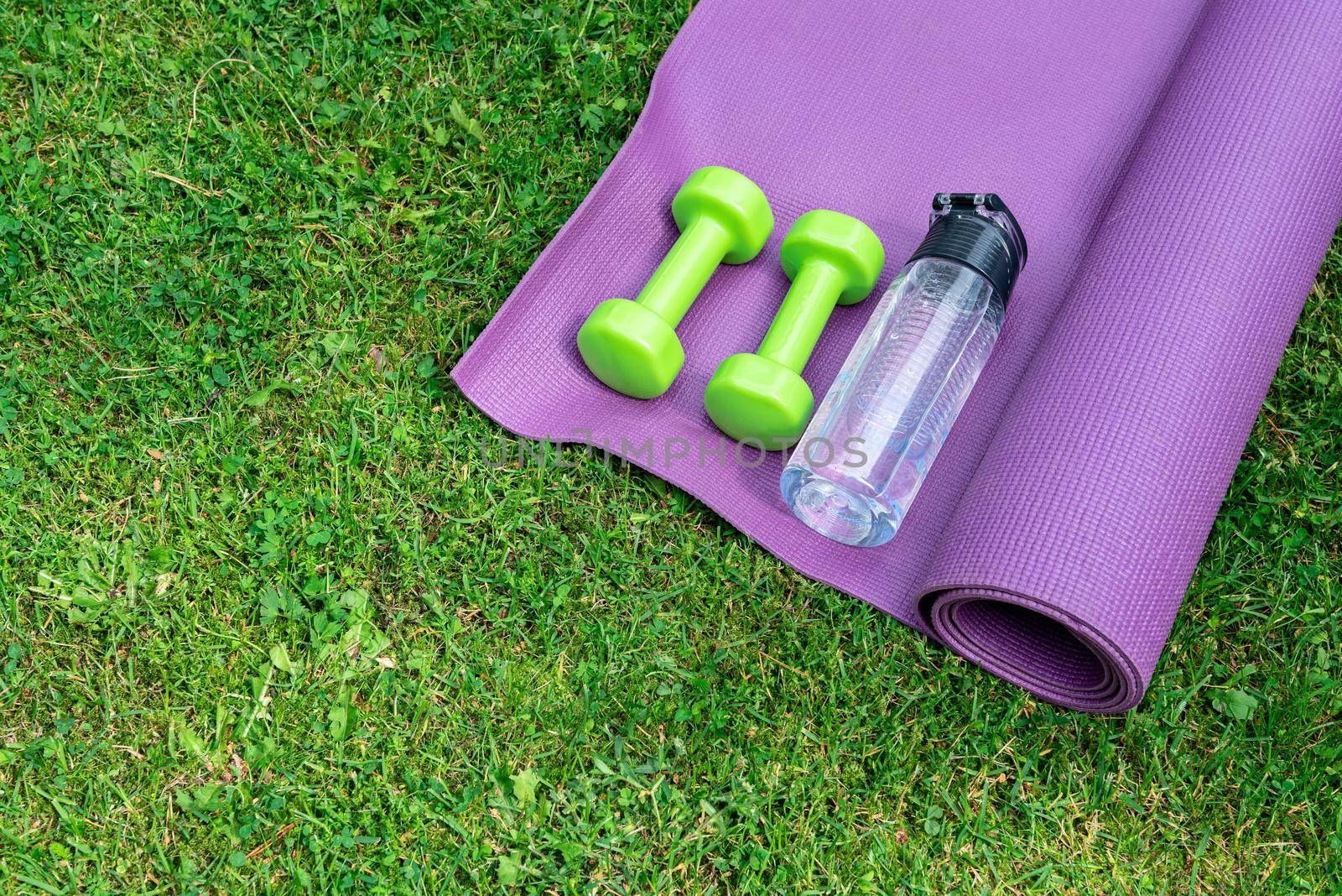 Ladie's dumbbells, water bottle and fitness mat on the green grass background, top view. Outdoor training concept. Copy-space.