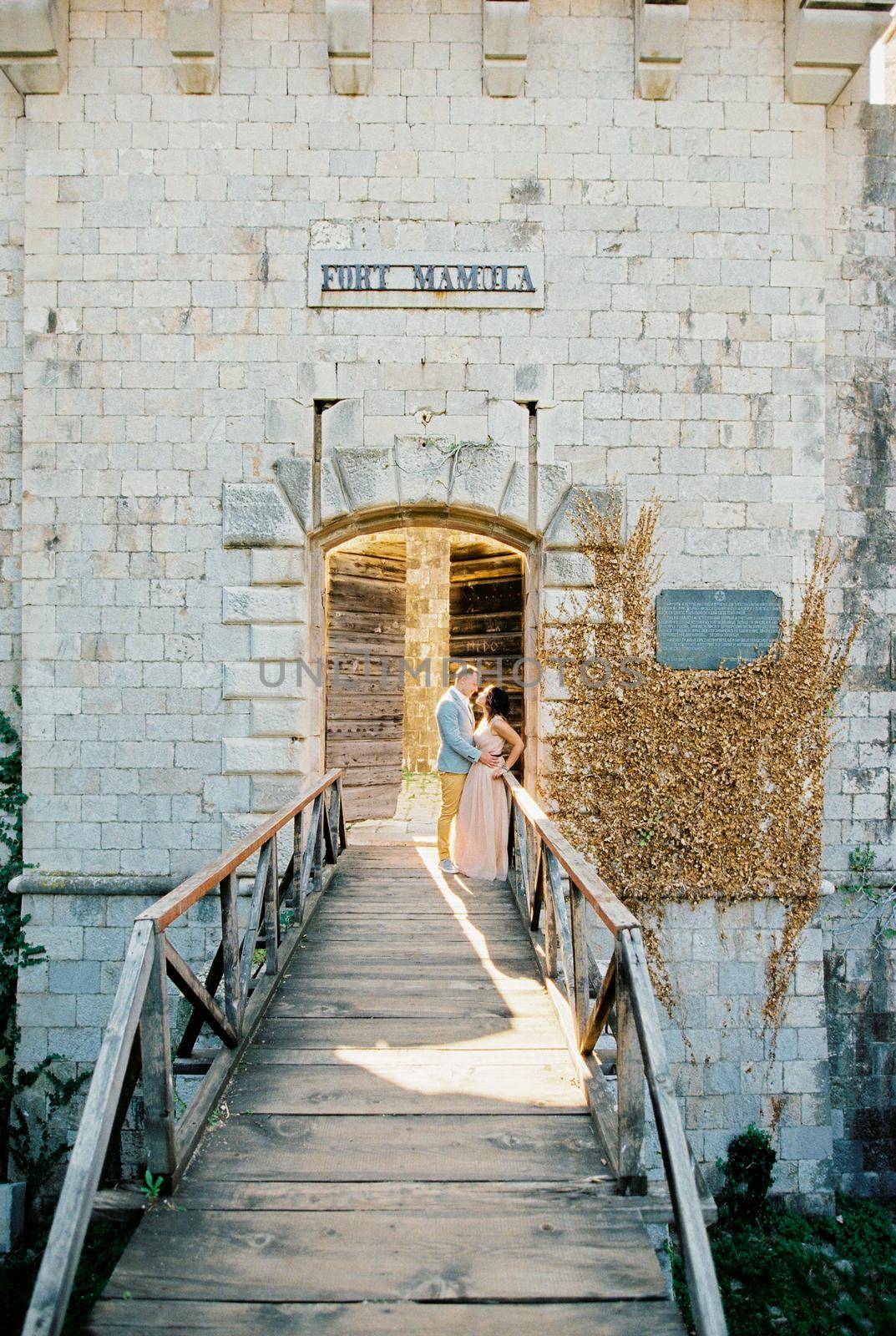 Groom hugs bride leaning against the railing of the wooden bridge to Mamula Fort. High quality photo