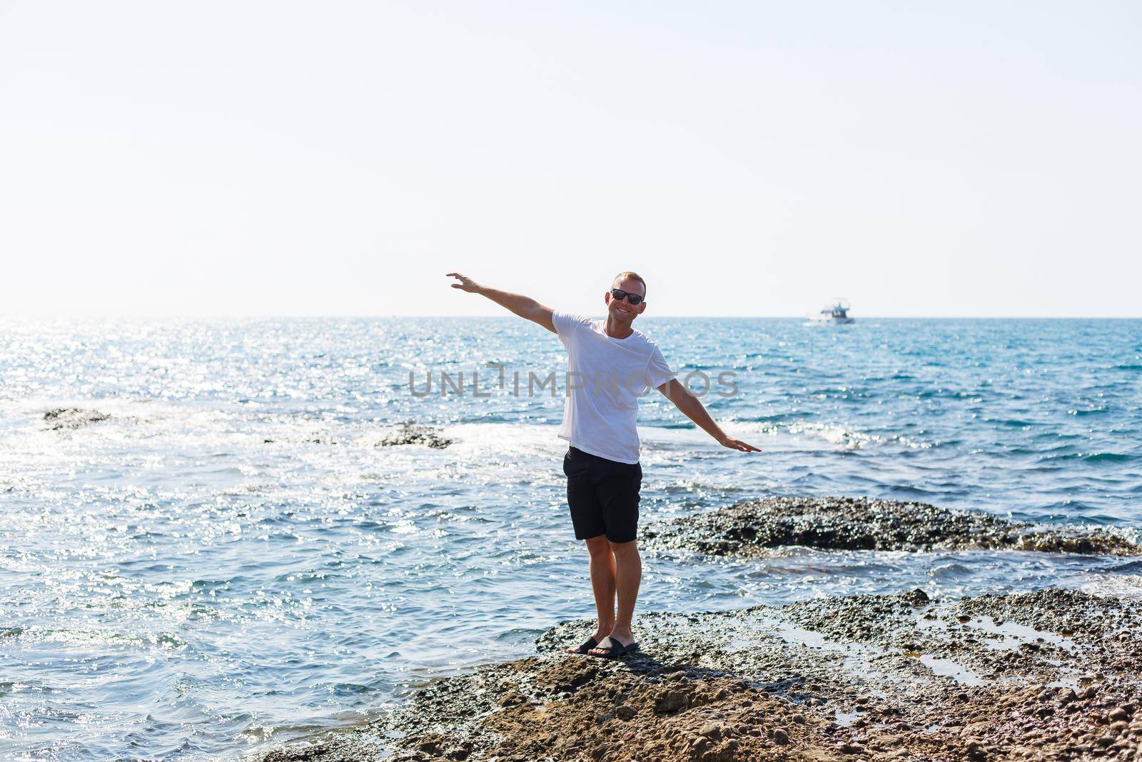 Young attractive man in sunglasses in a white t-shirt and shorts stands on the shore of the mediterranean sea by Dmitrytph
