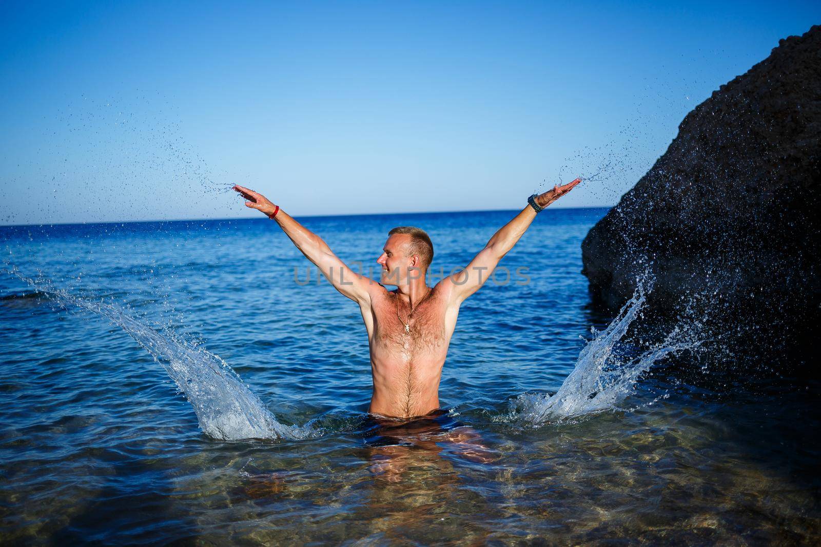 Happy man guy joy on vacation, stands at sea and splashes in the water. Men's shorts, sun, beach