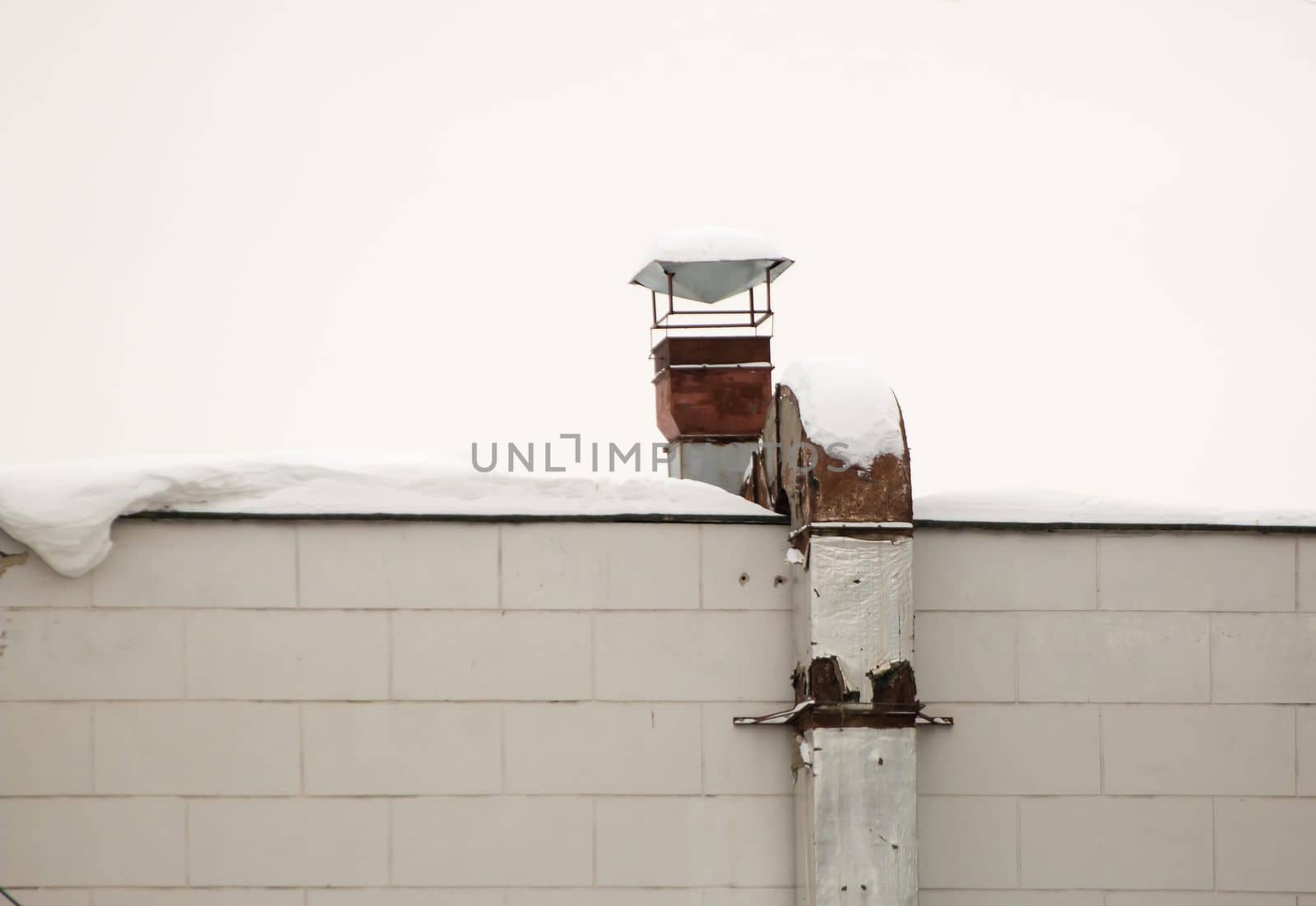 Metal rusty square pipe for exhaust and ventilation on the wall of the house. The roof of the building in white snow. Cloudy, cold winter day, soft light.