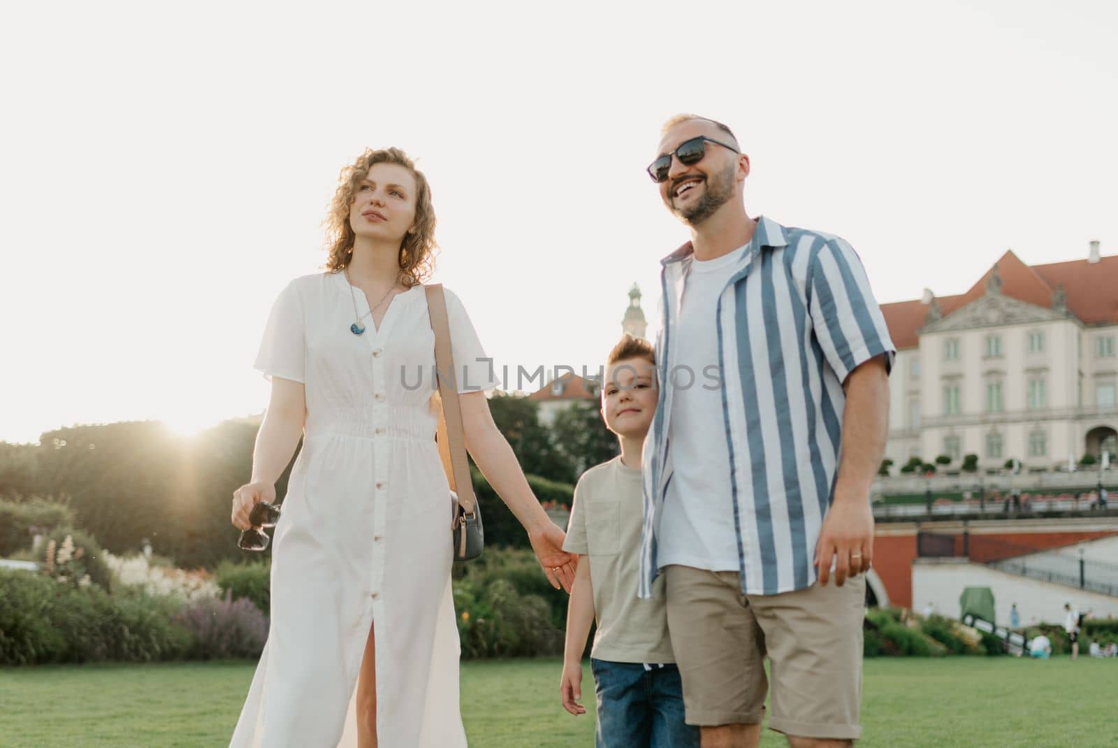 Father, mother and son are hugging in the garden of a European town. Happy family in the evening. Dad is laughing with his family in the background of the palace at sunset.