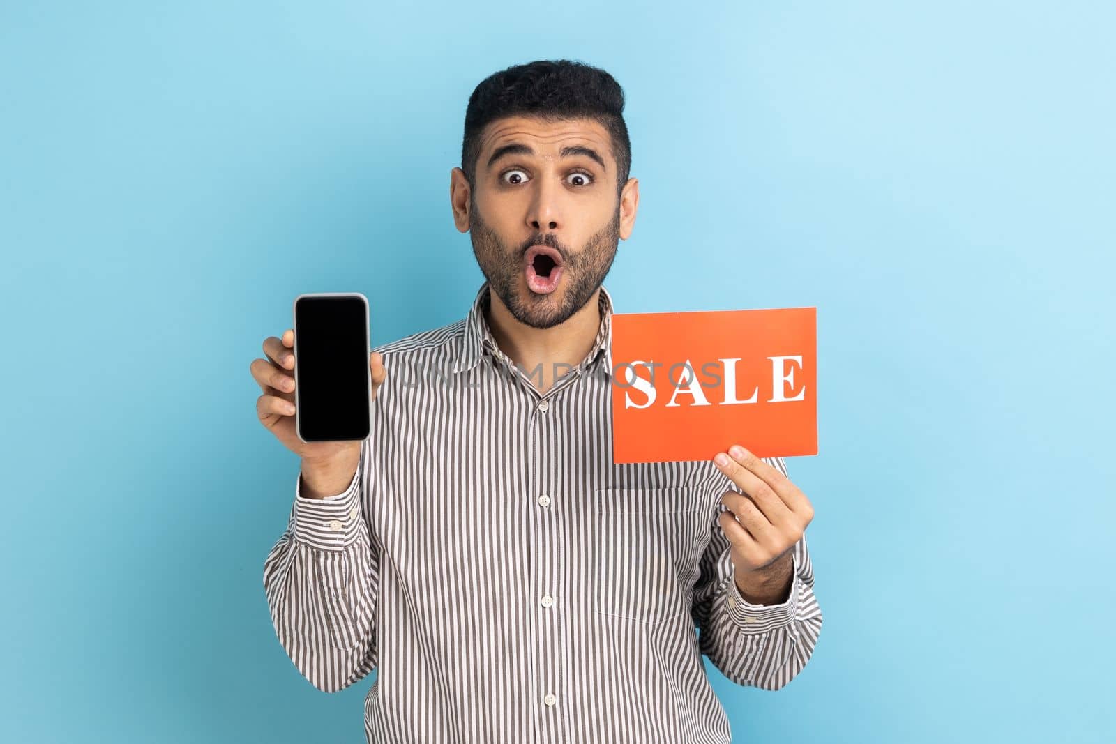 Amazed businessman holding sale inscription, cellphone and looking at camera with open mouth, showing discounts on mobile device, wearing striped shirt. Indoor studio shot isolated on blue background.