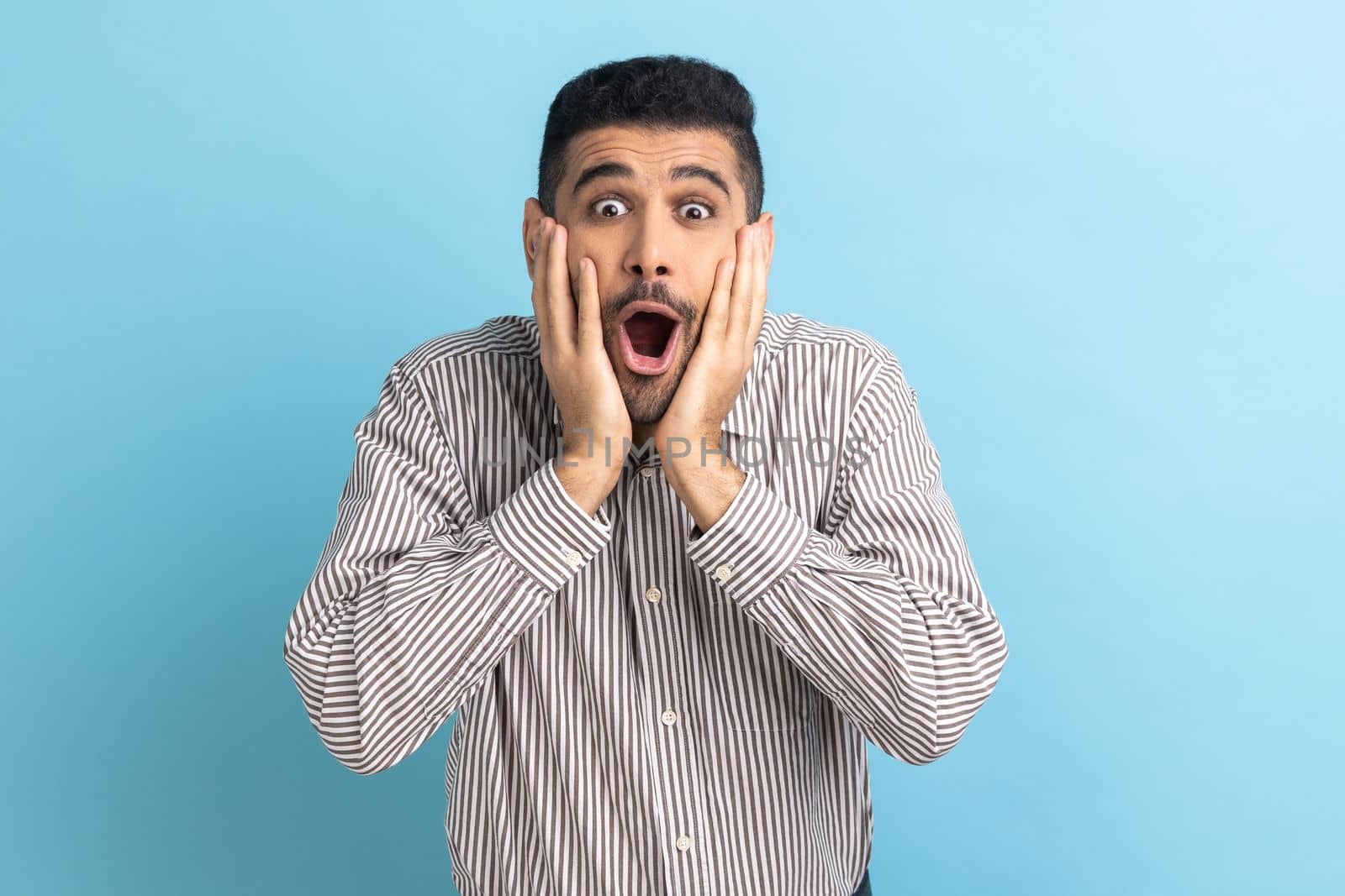 Surprised bearded businessman standing with open mouth and keeps hands on cheek, being impressed of shocking news, wearing striped shirt. Indoor studio shot isolated on blue background.